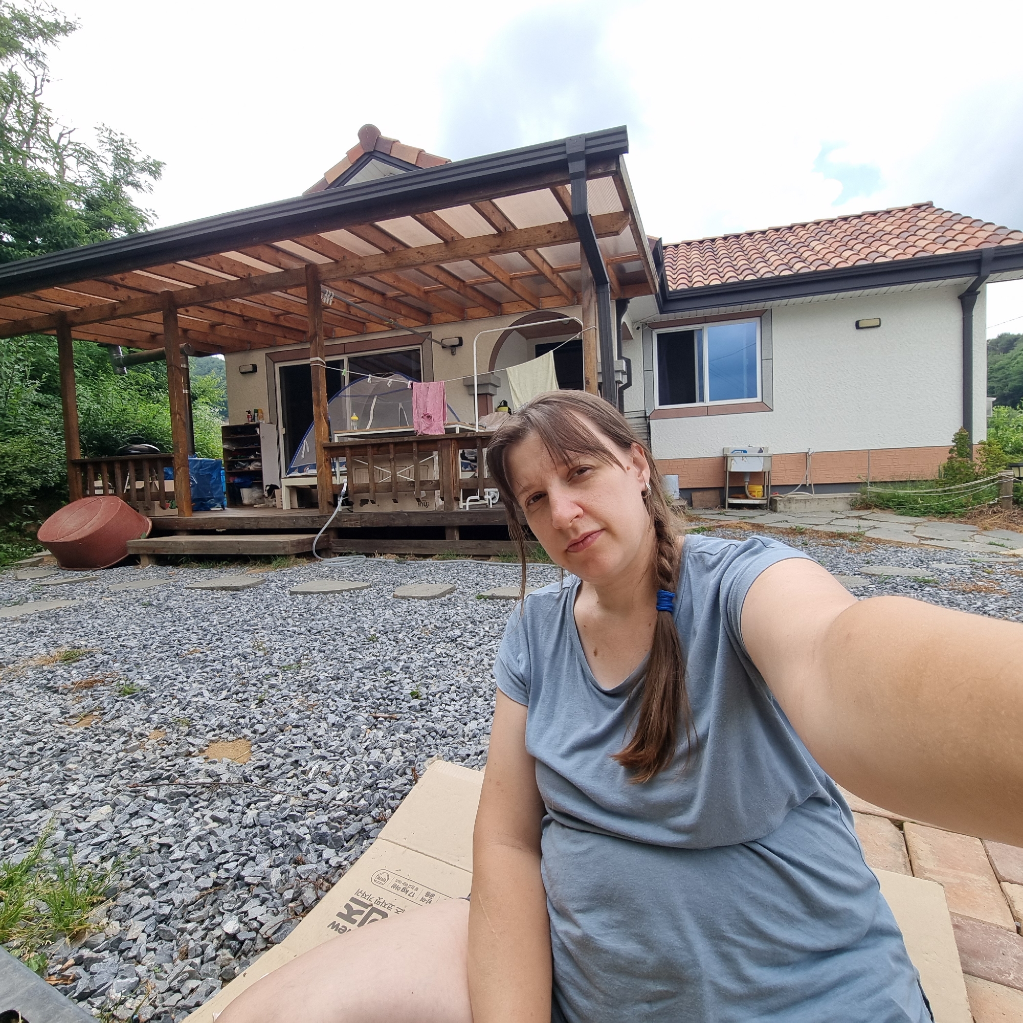 A woman with brown hair in a braid, wearing a blue t-shirt, takes a selfie in front of a house with a wooden porch and a tiled roof. The house appears to be in a rural or suburban setting, with gravel in the foreground and trees in the background. The woman is sitting on what appears to be a cardboard box, and her arm is extended towards the camera. There is a large red tub on the porch to her left, and laundry is hanging on a clothesline behind her. The location is Dunnae-myeon, South Korea.