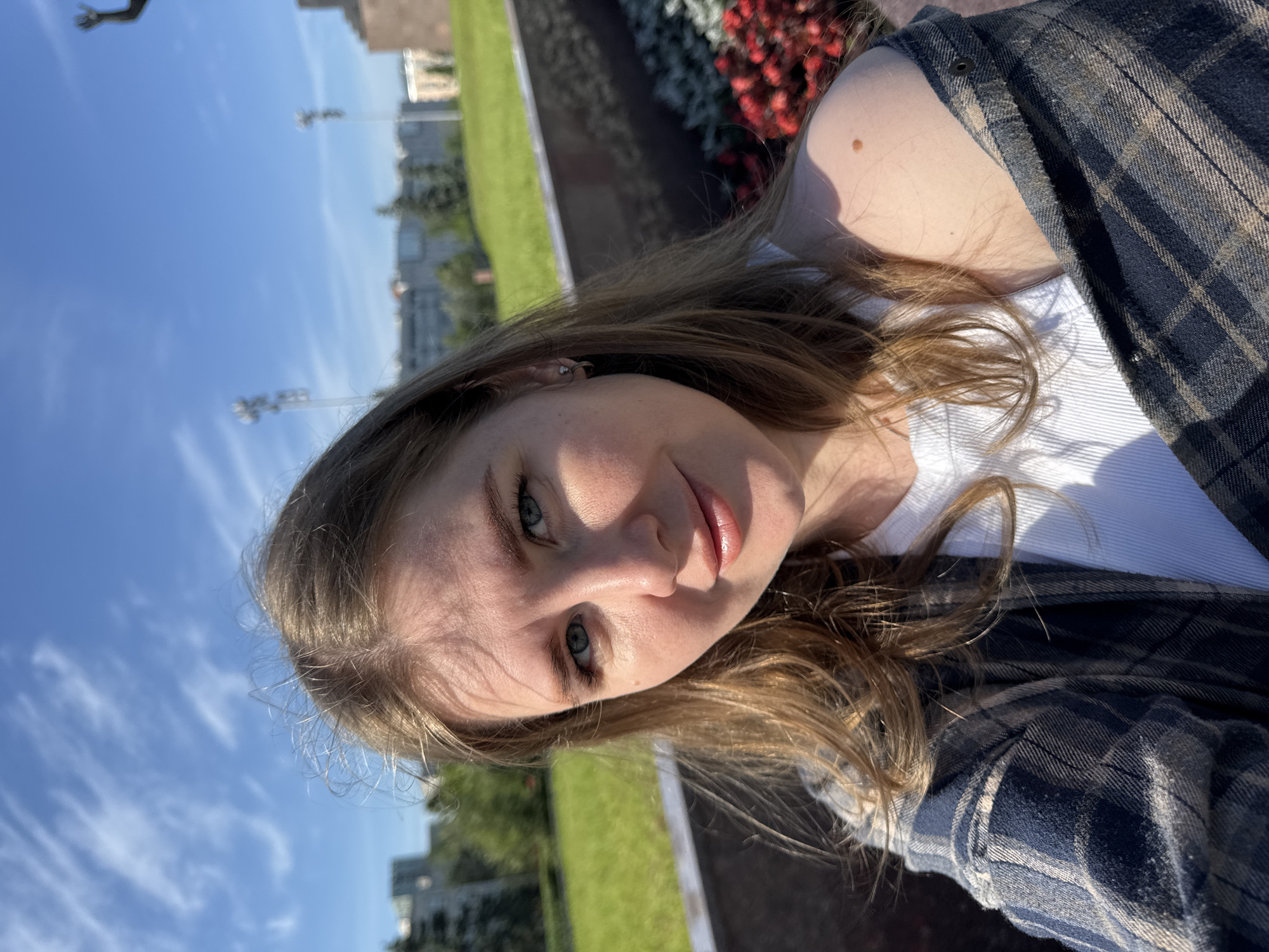 A young woman is featured in a close-up outdoor selfie, looking directly at the camera with a calm, subtle smile. She has long, wavy light brown hair, blue-green eyes, and fair skin, with sunlight illuminating one side of her face. She is wearing a white top under a dark blue and beige plaid shirt.
The background reveals a sunny park environment. A clear blue sky with scattered white clouds occupies the upper portion of the scene. Below, a vibrant green lawn is visible, bordered by a dark flower bed filled with red blossoms. In the distance, several buildings suggest an urban or suburban setting. A partial view of a large, indistinct monument or structure can be seen in the far background. The bright lighting indicates it is daytime. The location context provided is Perm, Russia. No text is visible in the image.