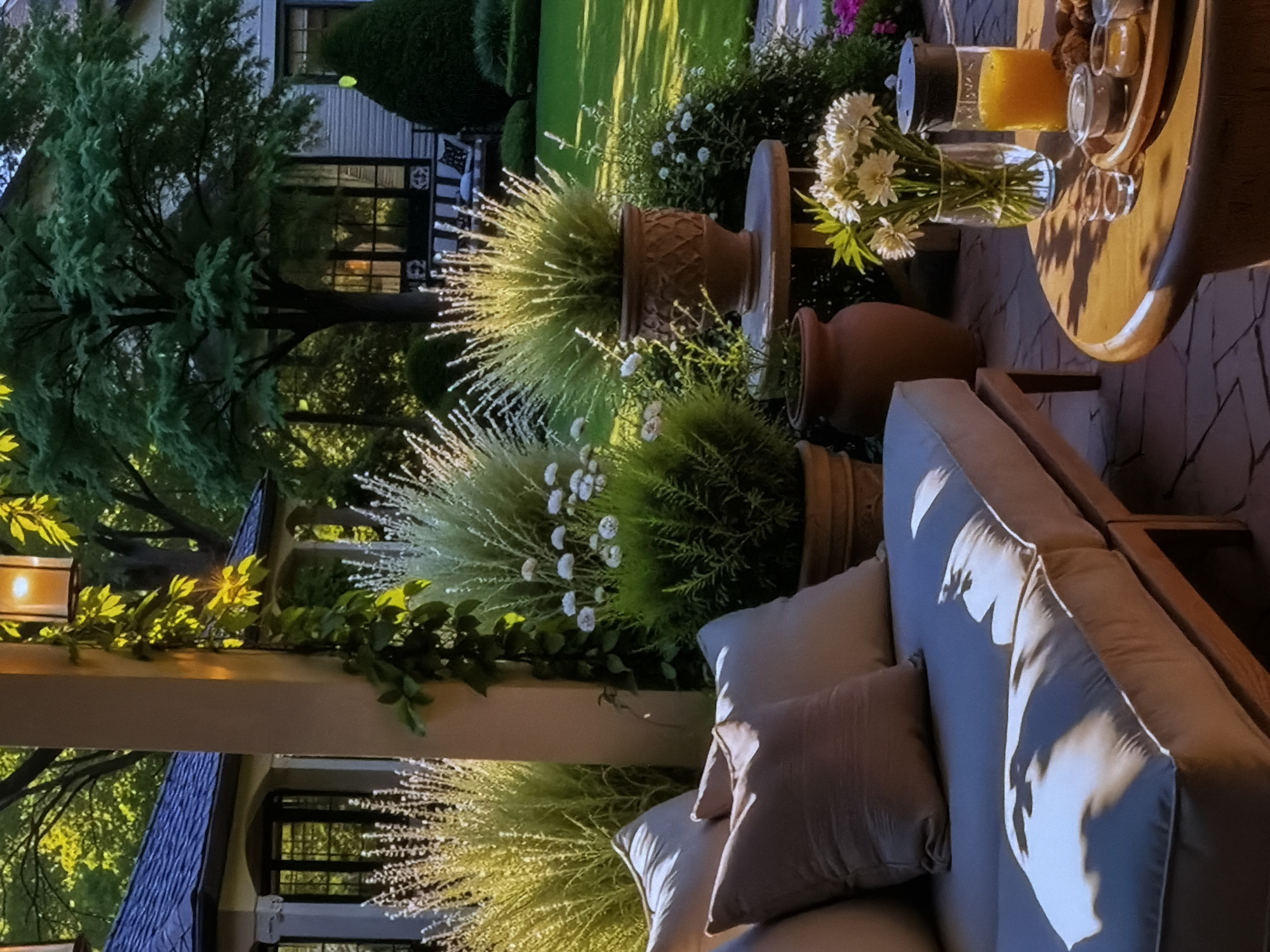 This image captures a serene and inviting outdoor patio setting, likely in a garden or backyard in Sofiivska Borshchahivka, Ukraine. No people are visible, suggesting a peaceful moment or a space prepared for future guests.
The scene is illuminated by warm, golden light, indicating either late afternoon or early evening. Long, distinct shadows are cast by objects like the table and foliage, contributing to a cozy and relaxed ambiance. A lantern with a glowing candle, visible on the left, further supports the evening setting.
In the foreground, a comfortable outdoor sofa with light-colored cushions and multiple throw pillows occupies the lower right portion of the frame. The cushions show soft shadows, highlighting their texture.
To the right, a round wooden table holds a clear glass pitcher filled with an orange beverage, a vase containing white flowers (resembling daisies or chrysanthemums), and a wooden tray with small glass containers, possibly holding snacks or condiments.
The middle ground features an array of lush potted plants, including tall, wispy ornamental grasses with white-tipped blades that catch the golden light brightly, and smaller green bushes adorned with delicate white flowers. These plants are arranged in decorative terracotta pots of varying sizes.
In the background, a house with dark-framed windows and a light-colored exterior is partially visible through dense foliage. A large, mature tree with abundant green leaves stands prominently on the left, its upper branches glowing with reflected sunlight. Manicured lawns and other garden elements complete the verdant setting.
The overall impression is one of tranquility and comfort, a perfect spot for unwinding. No visible text is present in the image.