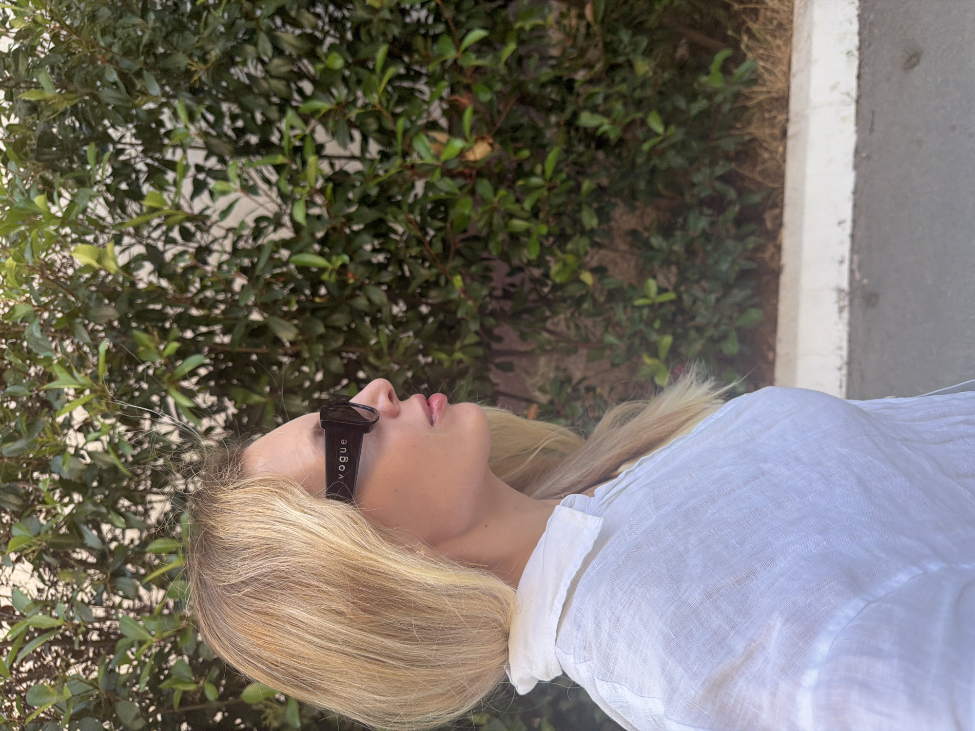 A blonde woman wearing a white linen shirt and black sunglasses with "Vogue" printed on them is shown in profile, facing right. She is standing outdoors in front of a dense green bush. The background also includes a hint of a white wall or fence and a sidewalk. The lighting suggests it is daytime. The woman's hair is shoulder-length and slightly windswept. Her facial features are visible, including her nose, lips, and cheek. The image is cropped to focus on the woman's upper body and head. The location is the city of Budva, Montenegro.