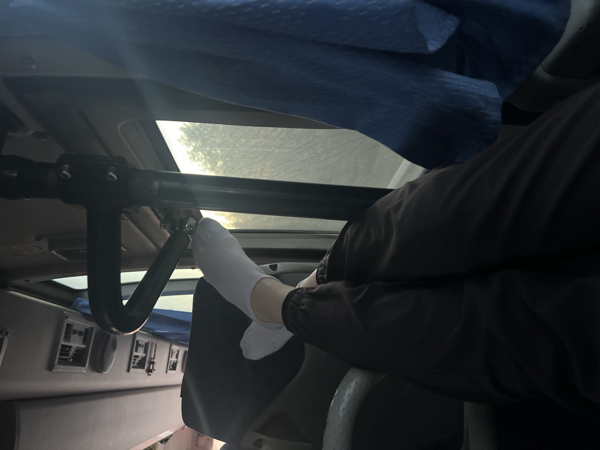 The photograph shows a view from inside what appears to be a vehicle, possibly a bus or van, with a person resting their legs on a seat. The person is wearing dark-colored pants and white socks. Part of the ceiling is visible, with a skylight and a black metal bar extending across the top. A blue cloth is draped above. On the side, there are a few air vents with control switches. The setting is unclear. There is no information about the city of Permyakova, Russia, in the photo.