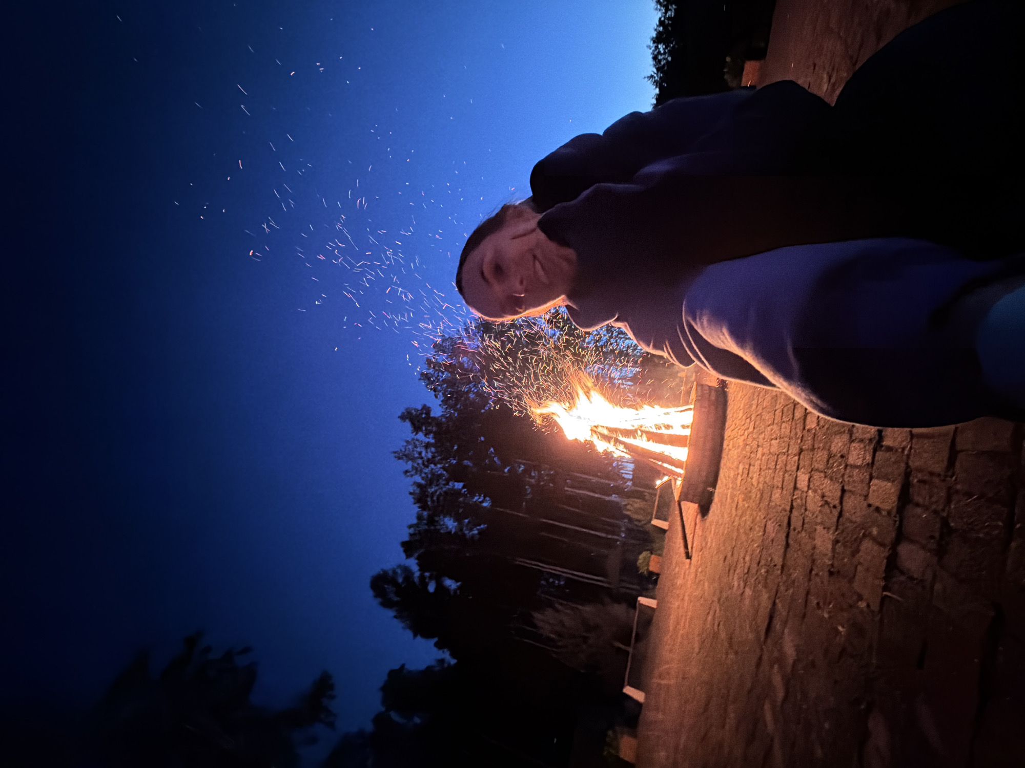 A young woman is pictured in a close-up selfie taken at dusk by a campfire. She is wearing a dark hooded sweatshirt. Her left hand is touching her cheek as she smiles at the camera. Behind her, a large bonfire burns brightly, sending sparks into the deep blue twilight sky. The fire is situated in a stone fire pit on a brick patio. In the background, silhouetted pine trees form a dark forest line. The overall scene suggests a recreational gathering outdoors. The location is identified as being in Birobidzhan, Russia.