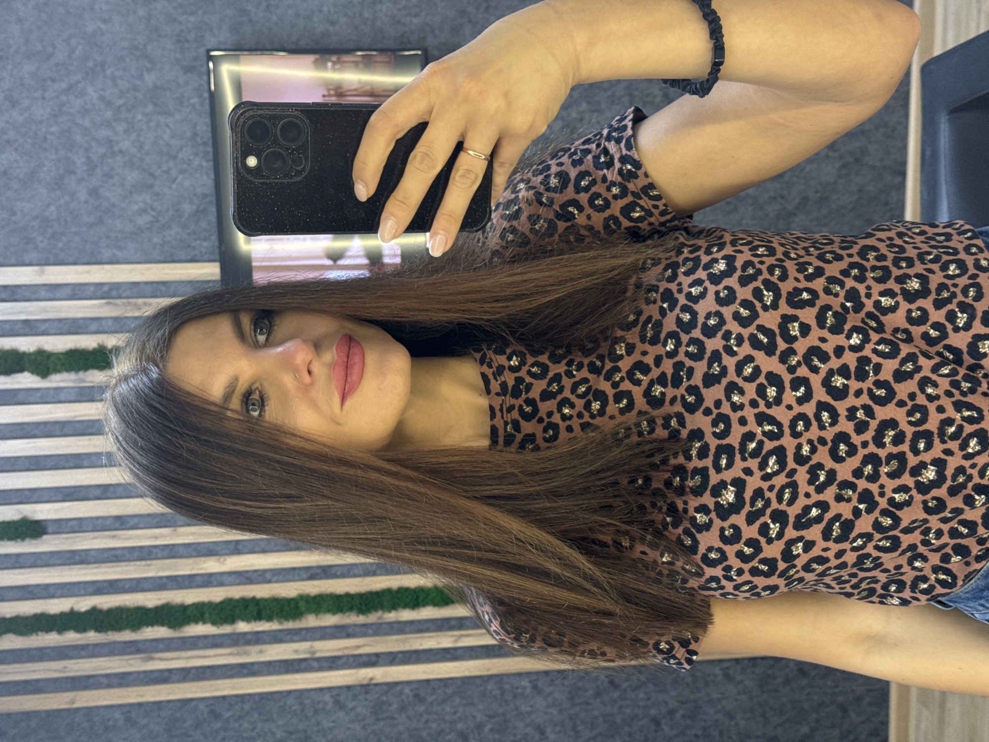 A woman is taking a selfie indoors. She has long brown hair and is wearing a brown leopard-print shirt. She is holding a black smartphone with multiple camera lenses. She has a gold ring on her finger and a black bracelet on her wrist. The background includes a gray carpeted wall, a decorative wall panel with horizontal stripes of wood and green moss, and a framed reflection that may contain artwork or lighting. There is no evidence within the image to confirm the location is in Chaikovsky, Russia.