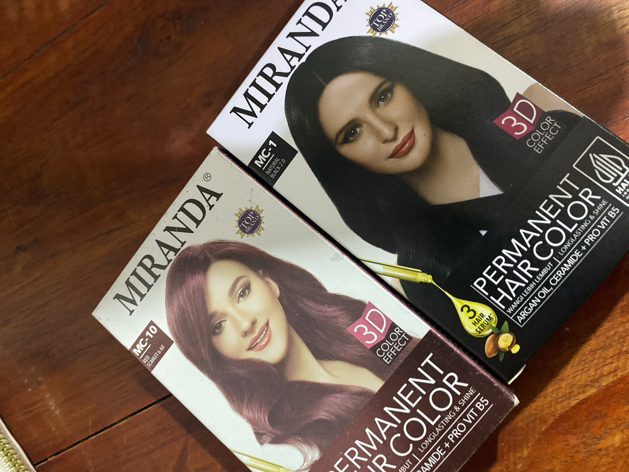 Two boxes of Miranda Permanent Hair Color are displayed on a wooden surface. The box on the left is lighter in color and features a woman with burgundy hair, along with text indicating "MC-10 RED SCARLET 6.60" and "3D COLOR EFFECT". The box on the right is darker and shows a woman with black hair, with text identifying it as "MC-1 NATURAL BLACK 2.0" and "3D COLOR EFFECT". Both boxes prominently display the "MIRANDA" brand name and a "TOP BRAND" seal. The scene appears to be indoors, possibly in a home environment in Ta', Indonesia, given the context provided.