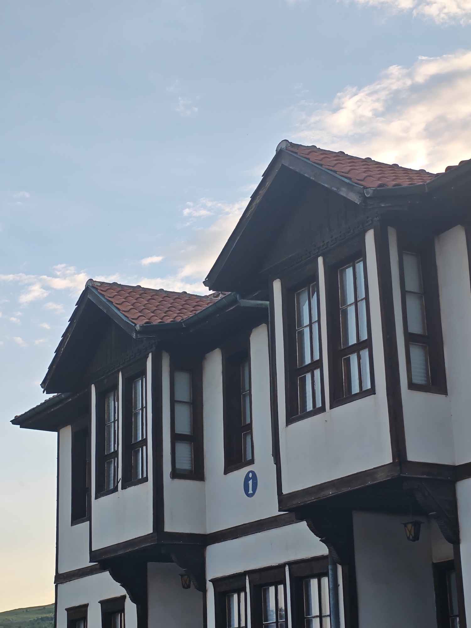 The image displays the upper sections of two adjacent traditional-style buildings located in Ivaylovgrad, Bulgaria, set against a partially cloudy, light blue sky. The architecture is characterized by white plaster walls contrasted with dark brown wooden beams, window frames, and structural supports. Both structures feature gabled roofs covered with reddish-brown terracotta tiles, arranged in an overlapping pattern. Dark wooden gutters are visible along the rooflines.
The upper floors of the buildings extend outward, supported by dark wooden corbels and beams. Multiple rectangular windows with dark brown frames are visible on the facades. Reflections of the sky are present in the windowpanes. One window on the rightmost building contains a partially visible white internal blind. Decorative carved patterns are discernible on some of the dark wooden eaves and under the roofline. A circular blue sign, bearing a white lowercase 'i' (indicating information), is affixed to the white wall of the left building, positioned below an upper-story window. In the lower-left background, a faint green, sloped landform is visible. The scene is illuminated by natural daylight; no individuals are present.