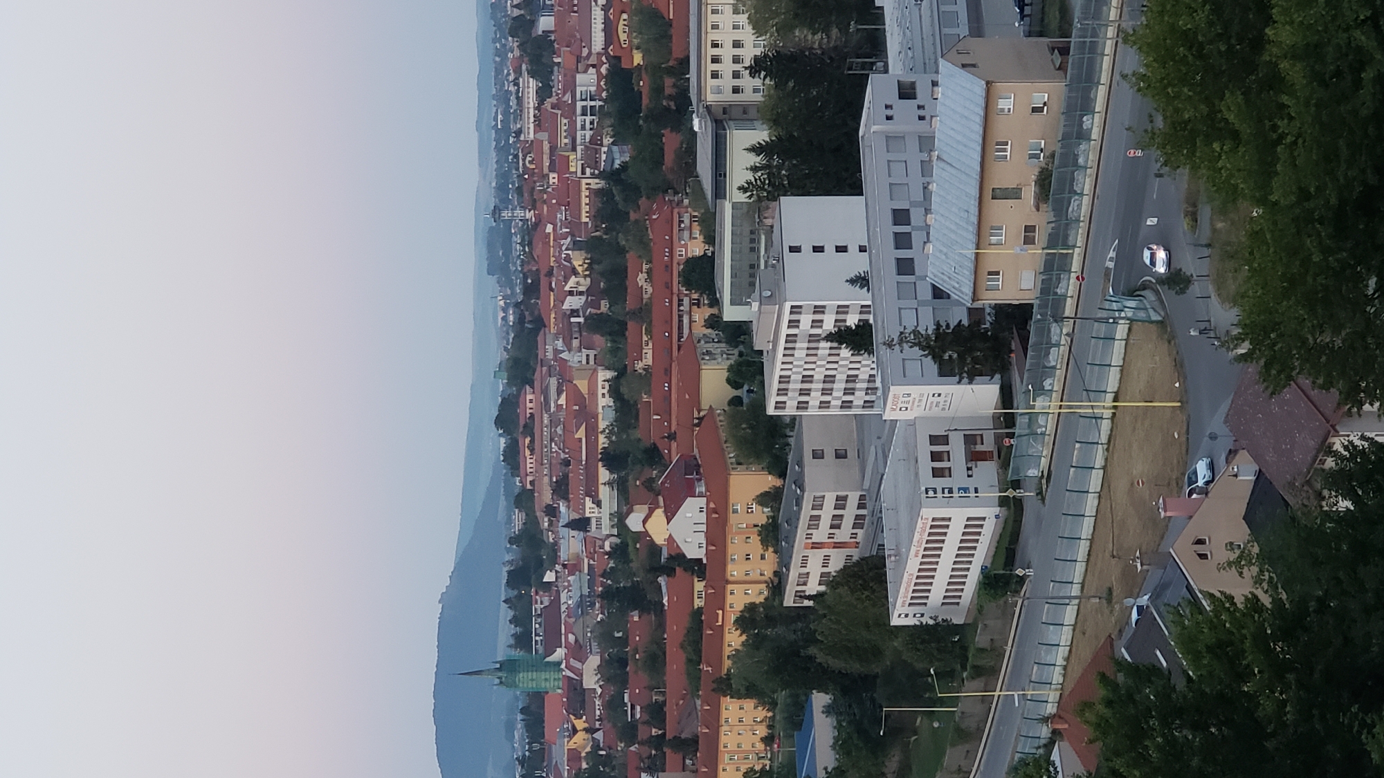 This is a panoramic aerial view of the city of Prešov, Slovakia, at dusk. The city is densely populated with buildings featuring red-tiled roofs, typical of European architecture. In the background, a large hill with some vegetation dominates the skyline, and a church steeple with a green spire is visible on its slope. In the foreground, there are several modern buildings with white and grey facades, a road with a sound barrier, and some trees. A few cars are seen on the road. The sky is a pale, gradient blue, indicating the fading light of the day. The overall scene depicts a residential and urban area transitioning into evening.