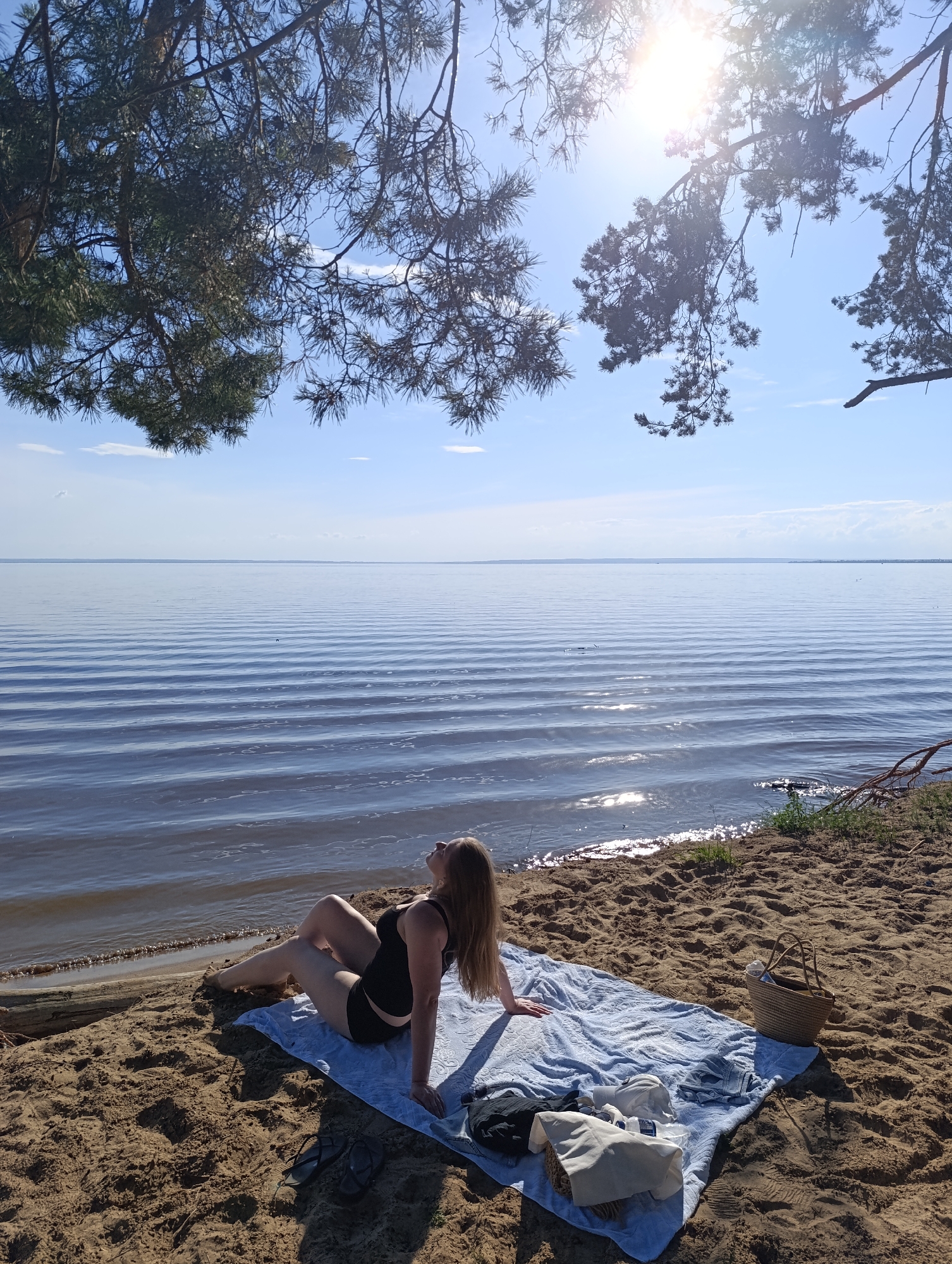 The image shows a woman sitting on a blue towel on a sandy beach, facing the water. She is wearing a black swimsuit and has her head tilted back, with her eyes closed, as if enjoying the sun. The beach is next to a calm body of water with gentle ripples. The sun is shining brightly in the sky, casting a glare on the water. There are pine trees hanging over the scene from above, with their branches casting shadows on the sand. Several items are placed on the towel next to the woman, including a straw bag, a white tote bag, some folded clothes, and a pair of flip-flops. The scene appears to be set during the daytime, likely in the late morning or early afternoon, given the angle of the sun. The overall mood of the image is peaceful and relaxing, suggesting a day spent enjoying the outdoors and nature. The location is not specified, but the presence of pine trees and a large body of water suggests a coastal or lakeside environment.