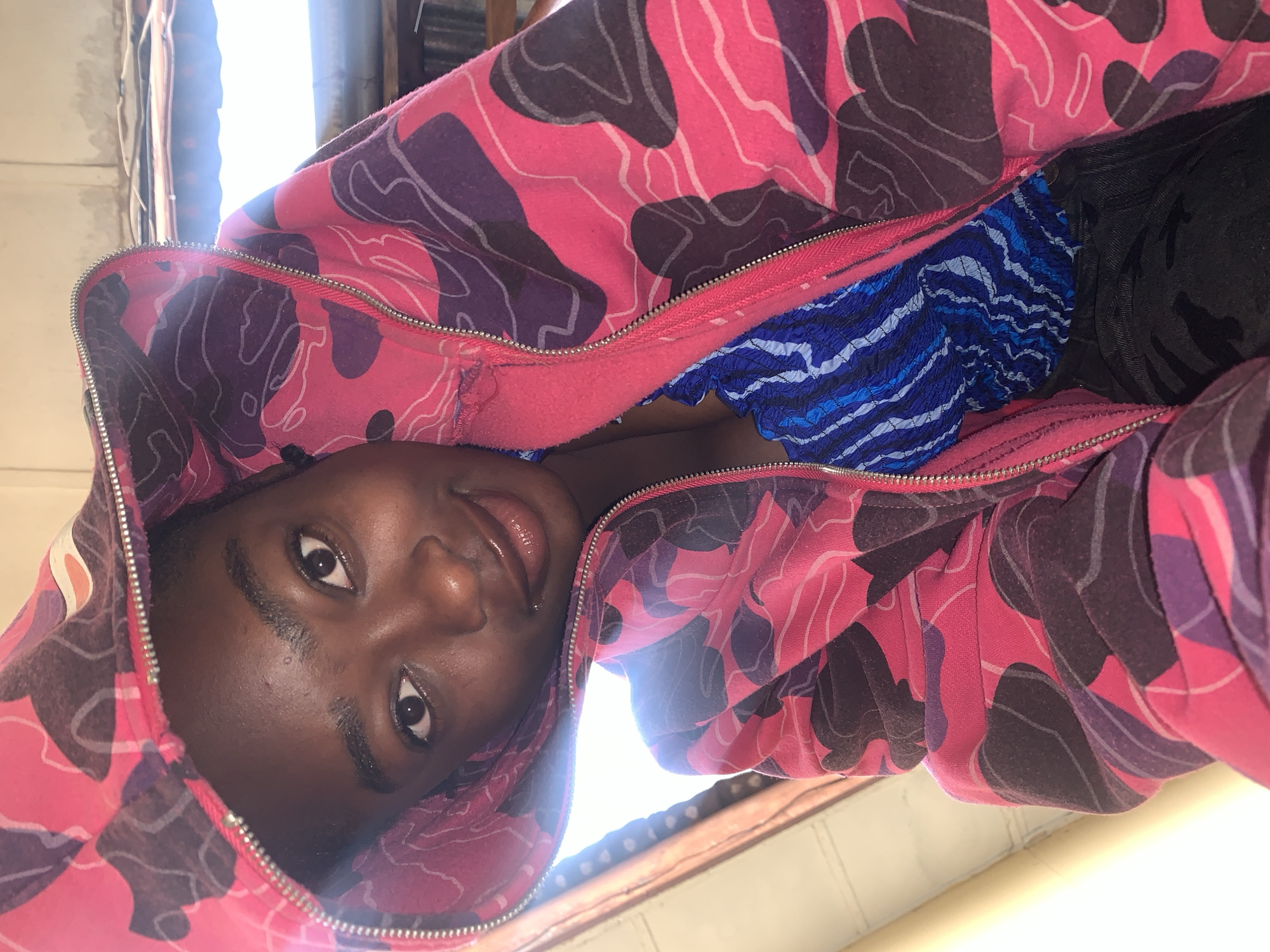 A young individual with dark skin is depicted in a close-up, eye-level selfie, looking directly at the camera with a subtle smile. The person wears a hooded, full-zip sweatshirt featuring a vibrant camouflage pattern dominated by bright pink, with irregular shapes in dark purple/maroon and thinner white outlines. The silver-toned zipper is partially open, revealing a blue top underneath, which has a textured, possibly smocked or shirred fabric with irregular light and dark blue stripes. The hood is up, framing the face. The individual has dark brown eyes, neatly shaped eyebrows, and a small metallic stud piercing visible on the left side of their upper lip.
The background suggests an indoor or sheltered setting in Rafin Par, Nigeria, with strong natural light entering from the upper left and upper right, causing some overexposure in those areas. Visible environmental details include a light-colored, possibly plastered or concrete wall with some staining or wear along the top edge in the upper right quadrant. A dark, possibly wooden beam or ledge is present below the wall. Further into the background on the right, what appears to be a corrugated metal structure or roofing is partially visible, with bright light filtering in. On the far left, a vertical light-colored structure, possibly a wall edge or doorframe, is present. The lower portion of the frame reveals dark-colored clothing, likely pants, with a faint pattern.