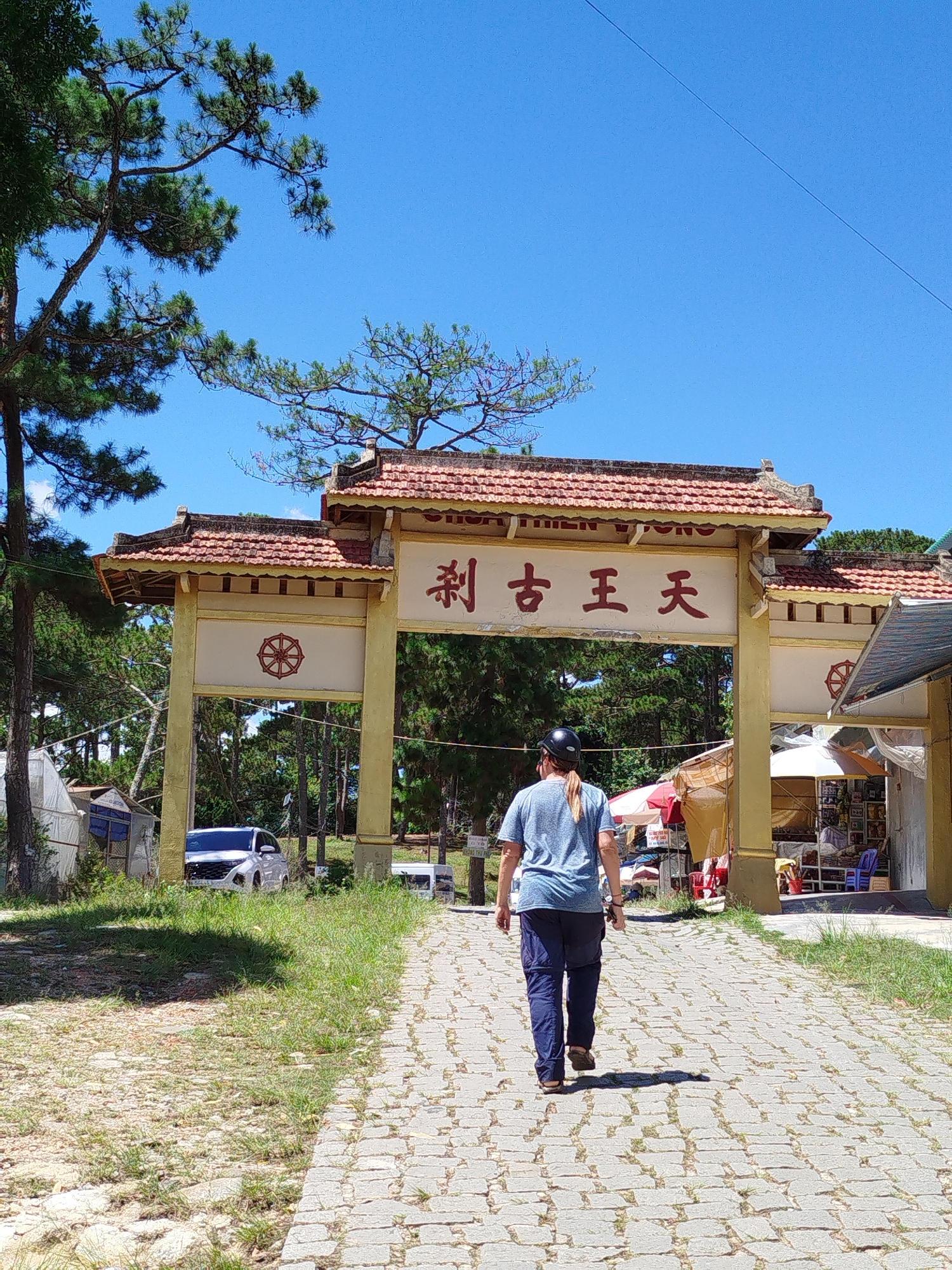 A sunny day view shows a person walking away from the camera towards a large yellow gateway. The gateway is tiled with red, and above the entrance are Chinese characters. To the left of the gateway is a car and to the right are several stalls selling goods. The person walking away is wearing a blue t-shirt and dark pants, sandals, and a black helmet. The gateway is on a gray brick road. Beyond the gateway are green pine trees under a clear blue sky. The location is Thien Vuong Co Sat Pagoda in the city of Da Lat, Vietnam.