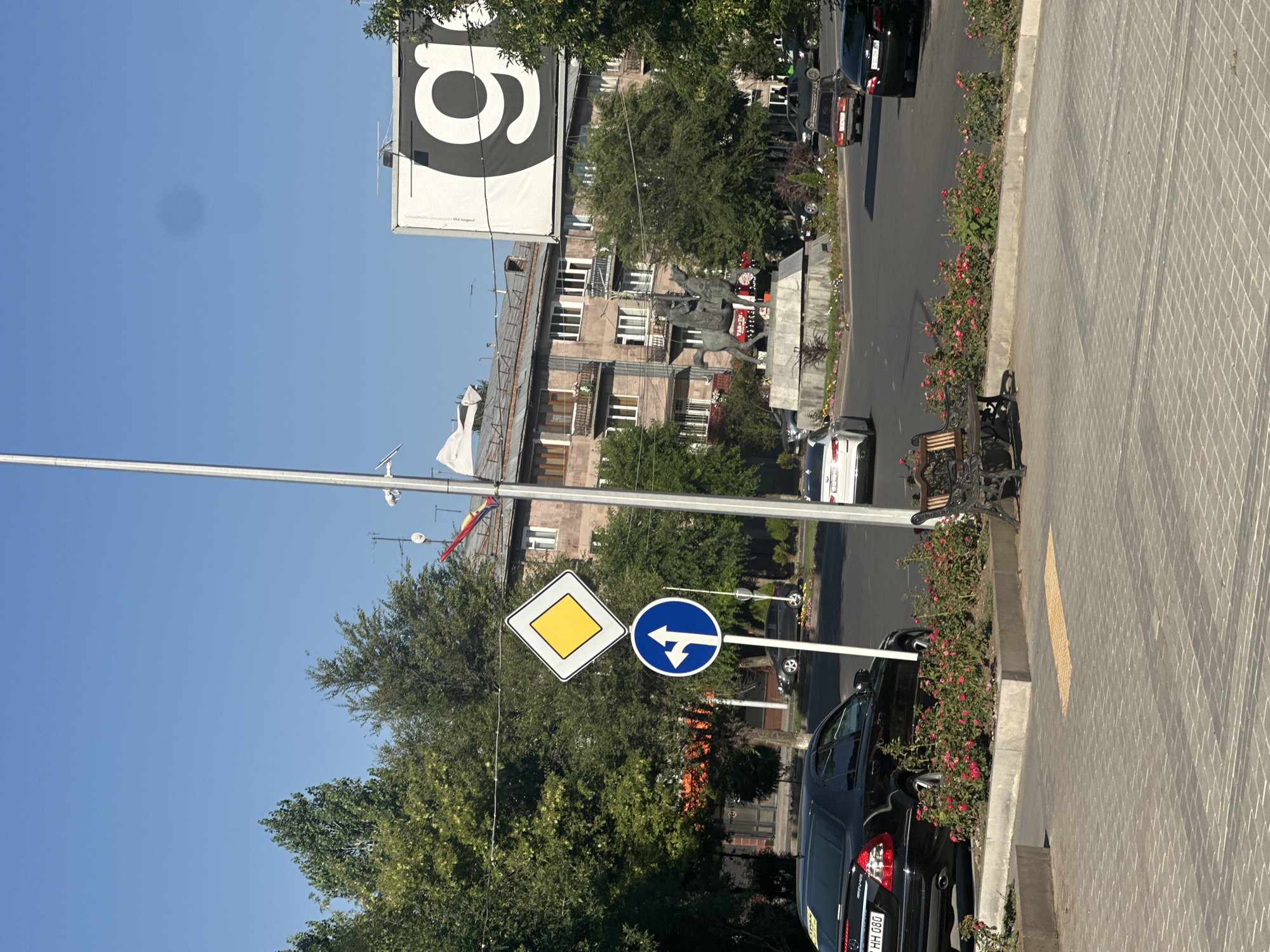 The image depicts a street scene in Gyumri, Armenia, under a clear blue sky. In the foreground, a paved walkway with a distinctive brick pattern is visible, featuring a tactile paving indicator and a metal bench with wooden slats. Adjacent to the walkway is a flowerbed filled with pink roses.
Dominating the middle ground is a traffic intersection. A black Mercedes-Benz sedan is parked on the left, its license plate partially visible. Traffic signs, including a diamond-shaped yield sign and a blue circular sign indicating a left turn and straight ahead option, are prominent. A tall, thin metal pole, likely a lamppost or utility pole, runs vertically through the center of the image.
Further into the scene, a statue of a man on horseback stands on a pedestal in the center of the intersection. Behind the statue, a multi-story building with several balconies and numerous windows is visible, showing architectural elements typical of Soviet-era construction. To the right of the building, a large billboard with a black "g" logo on a white background is prominently displayed. Several cars are navigating the intersection, and trees line the street, contributing to the urban environment. The overall impression is one of a bustling city intersection during daylight hours.
