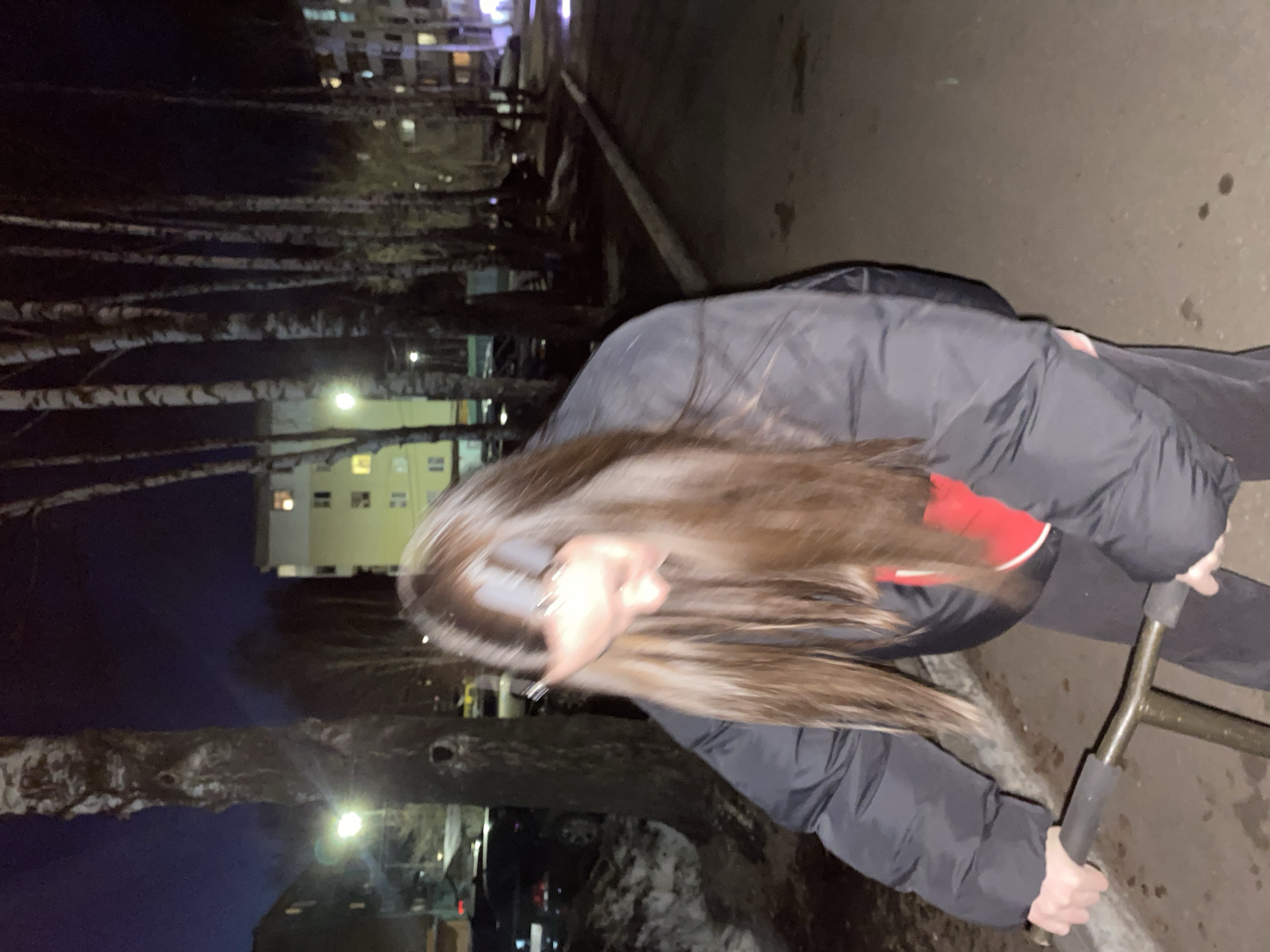 A young person with long brown hair wearing a dark jacket and red gloves is riding a scooter on a sidewalk. The person is in motion, causing a blur in the photograph. The location appears to be in the city of Votkinsk, Russia, evidenced by the typical apartment buildings and birch trees lining the street. It is nighttime, and streetlights are illuminating the area. Cars can be seen parked alongside the road.