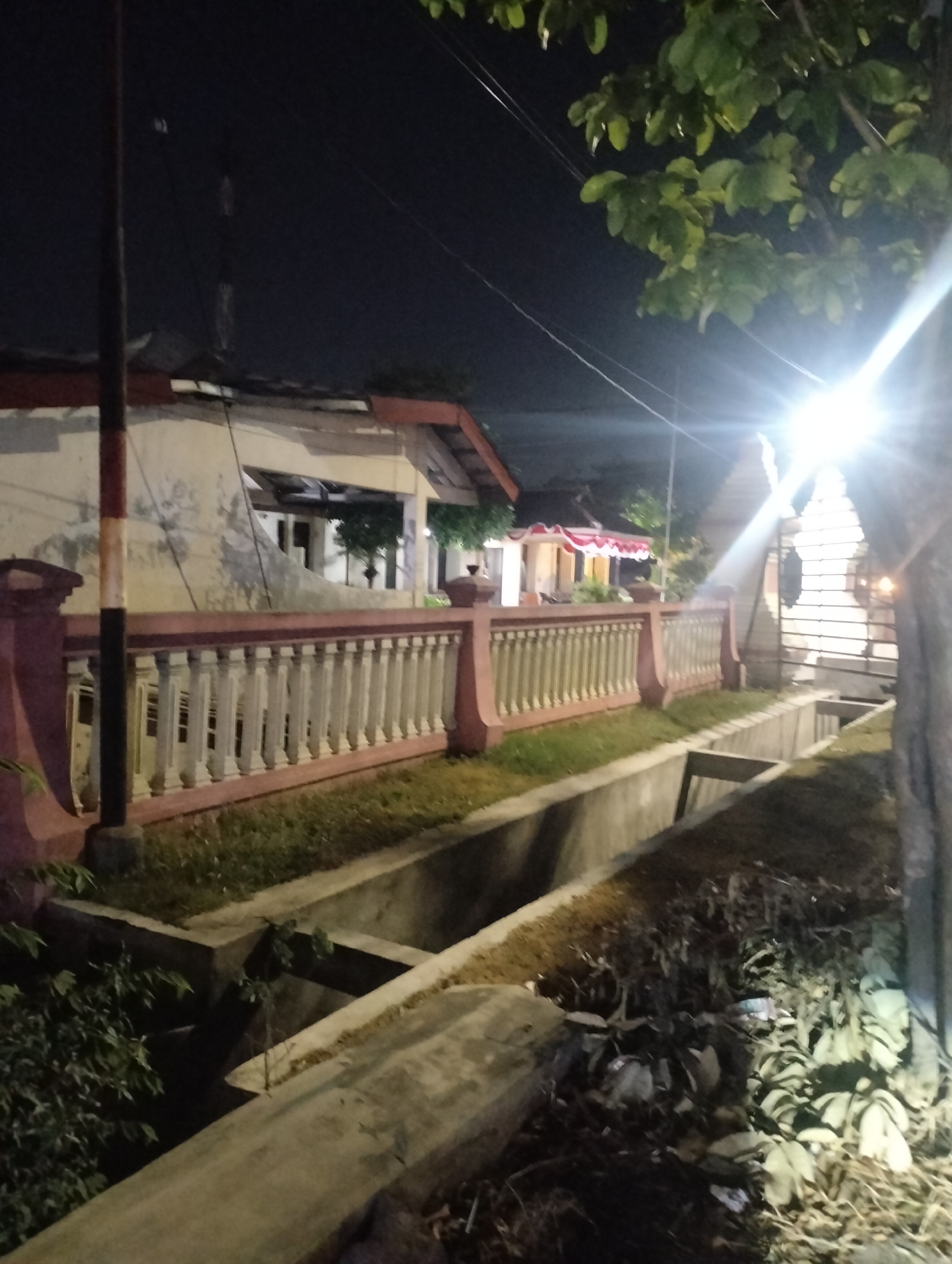 The image depicts a residential area in Driyorejo, Indonesia, at night. A concrete drainage channel runs horizontally across the foreground, bordered by overgrown vegetation on the left and dry grass and debris on the right. Behind the channel is a decorative fence with white balusters and a reddish-brown top railing. Beyond the fence, a building with a light-colored facade and a dark roof is visible, with a flagpole on the left side. Further down the street, another building with red and white awnings is illuminated. A bright light source, possibly a streetlamp, casts a strong glare on the right side of the image, illuminating tree leaves and a tree trunk. The overall scene is dimly lit by artificial light, suggesting it is nighttime. There are no people visible in the frame.