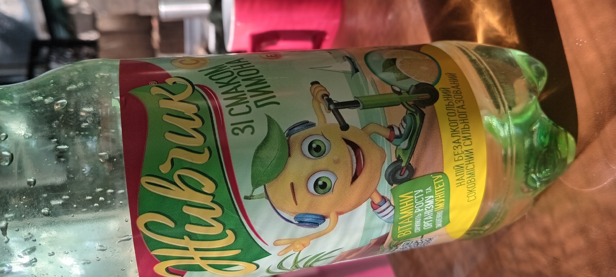 The image is a close-up, eye-level shot of a bottle of "Живчик" (Zhyvchyk) soft drink, a Ukrainian brand. The bottle has a green and yellow label featuring a cartoon lemon character wearing headphones and riding a scooter. Text on the label is in Ukrainian and reads "ЗІ СМАКОМ ЛИМОНА" (with lemon flavor), "ВІТАМІНИ СПРИЯЮТЬ РОСТУ ОРГАНІЗМУ ТА ЗМІЦНЕННЮ ІМУНІТЕТУ" (vitamins promote the growth of the organism and strengthen immunity), and "НАПІЙ БЕЗАЛКОГОЛЬНИЙ СОКОВМІСНИЙ СИЛЬНОГАЗОВАНИЙ" (non-alcoholic juice-containing highly carbonated drink). The bottle is placed on a wooden surface, likely a table, outdoors, with blurred background elements that suggest a sunny day. In the background, part of a red mug and some vegetation are visible. The presence of condensation on the bottle indicates it is a cold beverage. The image does not contain any information about the city of Koviagi, Ukraine, or any specific locations within it.