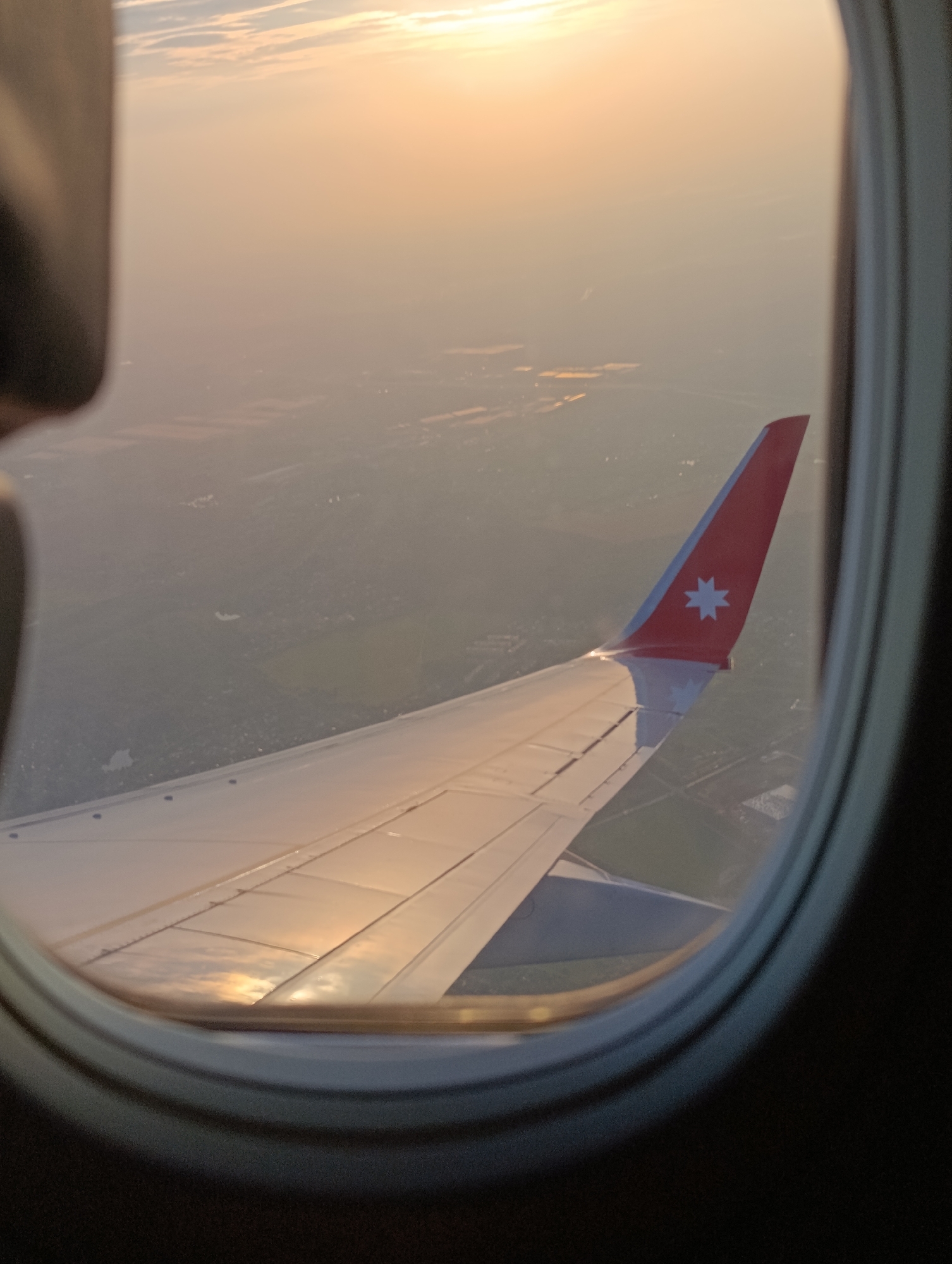 The image shows an airplane wing and tail as seen from a window seat. The wing is illuminated by the setting sun, casting warm reflections. The tail is red with a white star emblem. Below, a hazy landscape of fields and buildings is visible, suggesting the plane is flying over a rural area. The image is taken during golden hour, with the sky displaying soft orange and yellow hues. The photo has a slightly blurry quality, typical of a shot taken from an airplane window. There is no discernible information about Staroe Mart'yanovo, Russia in the provided image.