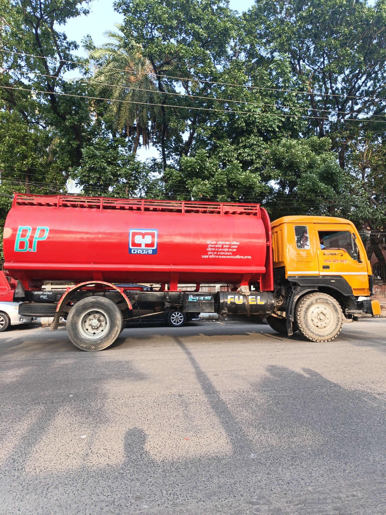 A bright, sunlit, full-shot photograph captures a red and yellow fuel tanker truck parked on an asphalt road, likely in an urban or semi-urban area. The truck dominates the central and lower portions of the frame. The tanker section is a prominent red cylinder with "B.P" in large teal letters on its side. Next to "B.P" is a square logo with "মেঘনা" (Meghna in Bengali) written below it. Further Bengali text, appearing to be contact or product details, is visible on the red tank: "ছিঁচকে নইলের এইঙন অধিপর", "দেখন বাগিন ঢাকা-১০০০", "নাইলের-১৯০২০০-০০১০", "মেনু-৮০০২৫১২", and "মরার সার্ভিস ৫-৫০১৭৩০-০০২০০১ ােজিগঞ্জ, রাগঞ্জ".
The truck's cab is bright yellow, and the brand "EICHER" is visible on its side, along with Bengali text "আমম ভরাহ আমান" (Our Trust Aman) and "আমারে পরিক্ষার". A small box below the cab is labeled "টুলবক্স" (Toolbox). Large white letters spelling "FUEL" are painted on the black undercarriage of the tank, near the cab. The truck has large, rugged tires.
Overhead, lush green trees fill the upper portion of the image, with glimpses of a clear, light blue sky. A palm tree is also visible amidst the dense foliage. Power lines stretch horizontally across the sky, passing through the trees. Shadows of the truck and what appear to be people or other objects are cast on the road, suggesting a clear, sunny day, possibly morning or late afternoon. A small part of a white car is visible behind the tanker's rear wheels.