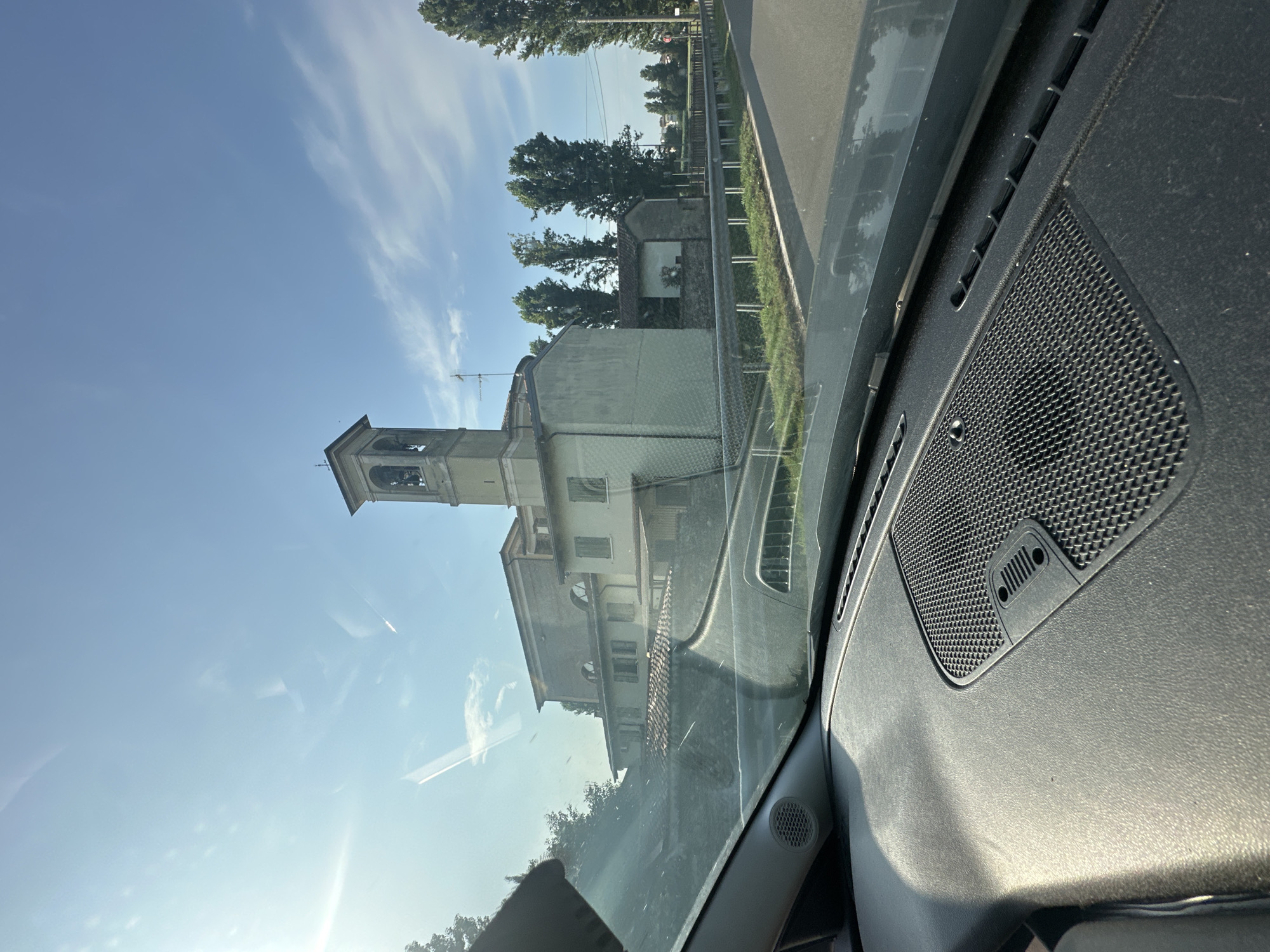 The image, taken from inside a vehicle, shows a view through the windshield of a building with a bell tower. The building appears to be a church or a similar religious structure, with a light-colored facade and a tiled roof. It is situated on the right side of the frame, with tall trees flanking it. A road is visible in the foreground, with a metal guardrail separating it from a grassy area. The sky is blue with scattered clouds, and the sun is shining, casting a bright light on the scene. The dashboard of the car is visible at the bottom of the frame, with a speaker grille prominent in the lower right quadrant. The image was captured in Pumenengo, Italy.