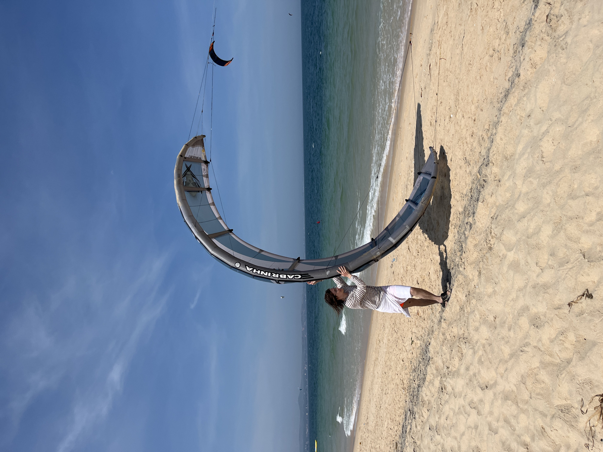 A young woman with brown hair is standing on a sandy beach in Phu Thuy Ward, Vietnam, holding a large, crescent-shaped kite. She is wearing a striped long-sleeved shirt and a white wrap around her waist. The kite is gray and white with the brand name "CABRINHA" visible. The ocean is a deep blue with white waves crashing on the shore. The sky is clear blue with some wispy clouds. In the distance, other kitesurfers can be seen in the water, along with a coastline and hills. The sand is light brown and appears to be slightly wet near the water's edge.