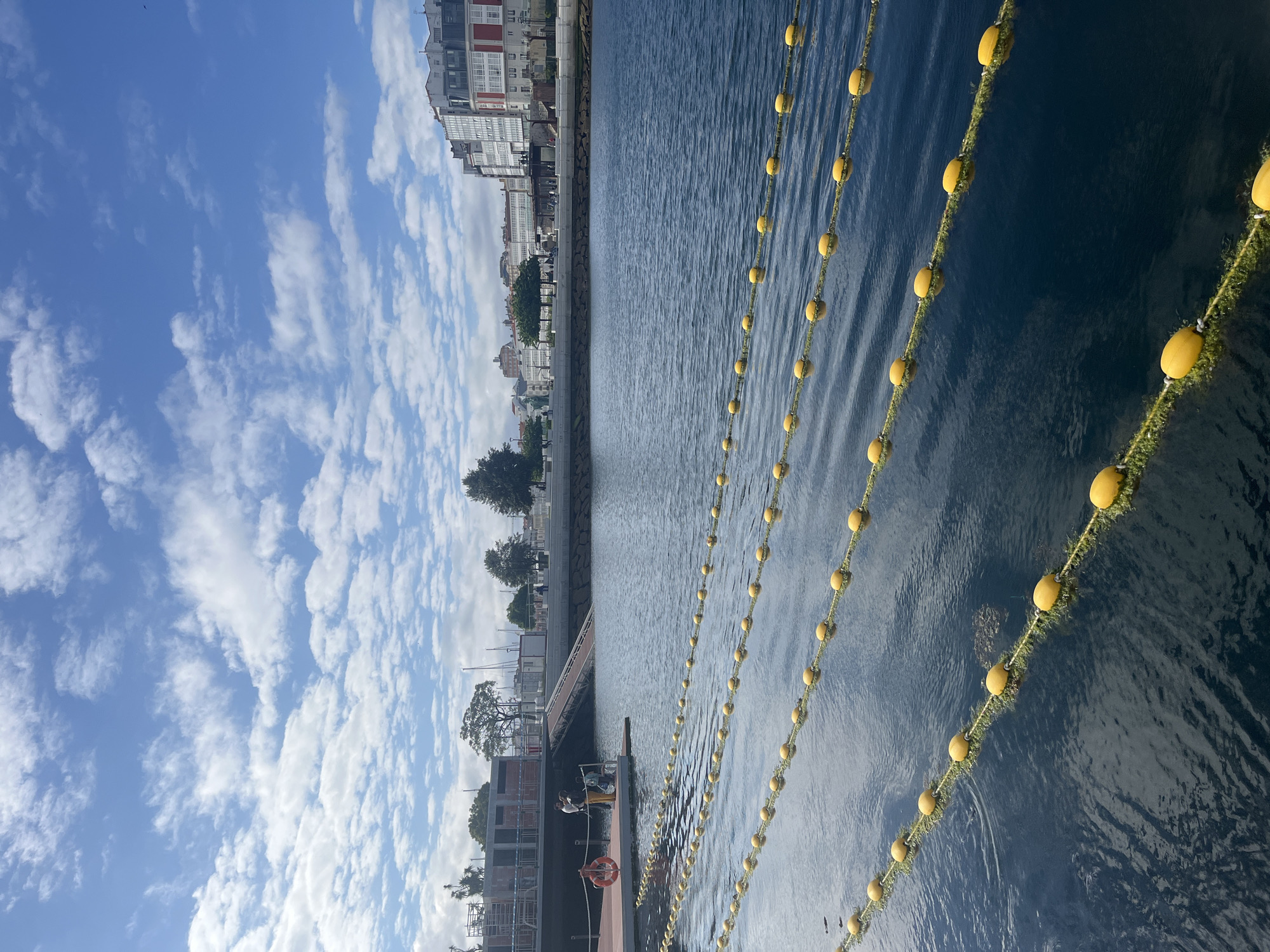 The image depicts a waterfront scene in A Coruña, Spain. The scene is characterized by a body of water with several rows of yellow buoys. A city skyline with apartment buildings is visible in the background. The sky is blue and partly cloudy. There are trees and possibly other foliage along the waterfront. A dock with a life ring, construction equipment, and people working are visible.
