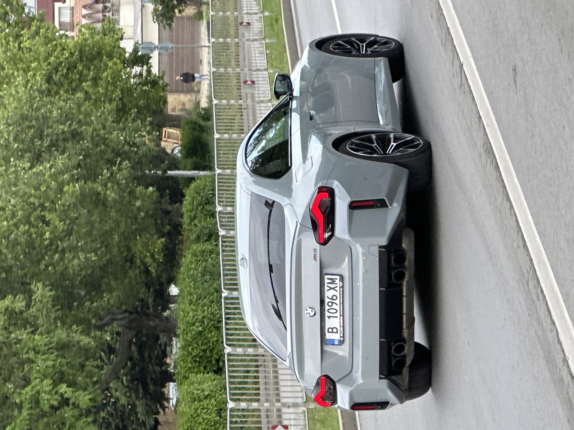 The image shows the rear view of a gray BMW M2 car with license plate "B 1096 XM". The car is parked on a gray asphalt road with a white line marking the lane. To the left of the car, there's greenery consisting of trees and hedges, as well as a metal fence. In the background, there's a street with buildings and a pedestrian walking on the sidewalk. According to the plate the car is located in Bulgaria. The car's details (model, plate) identify Varna as a very probable location.