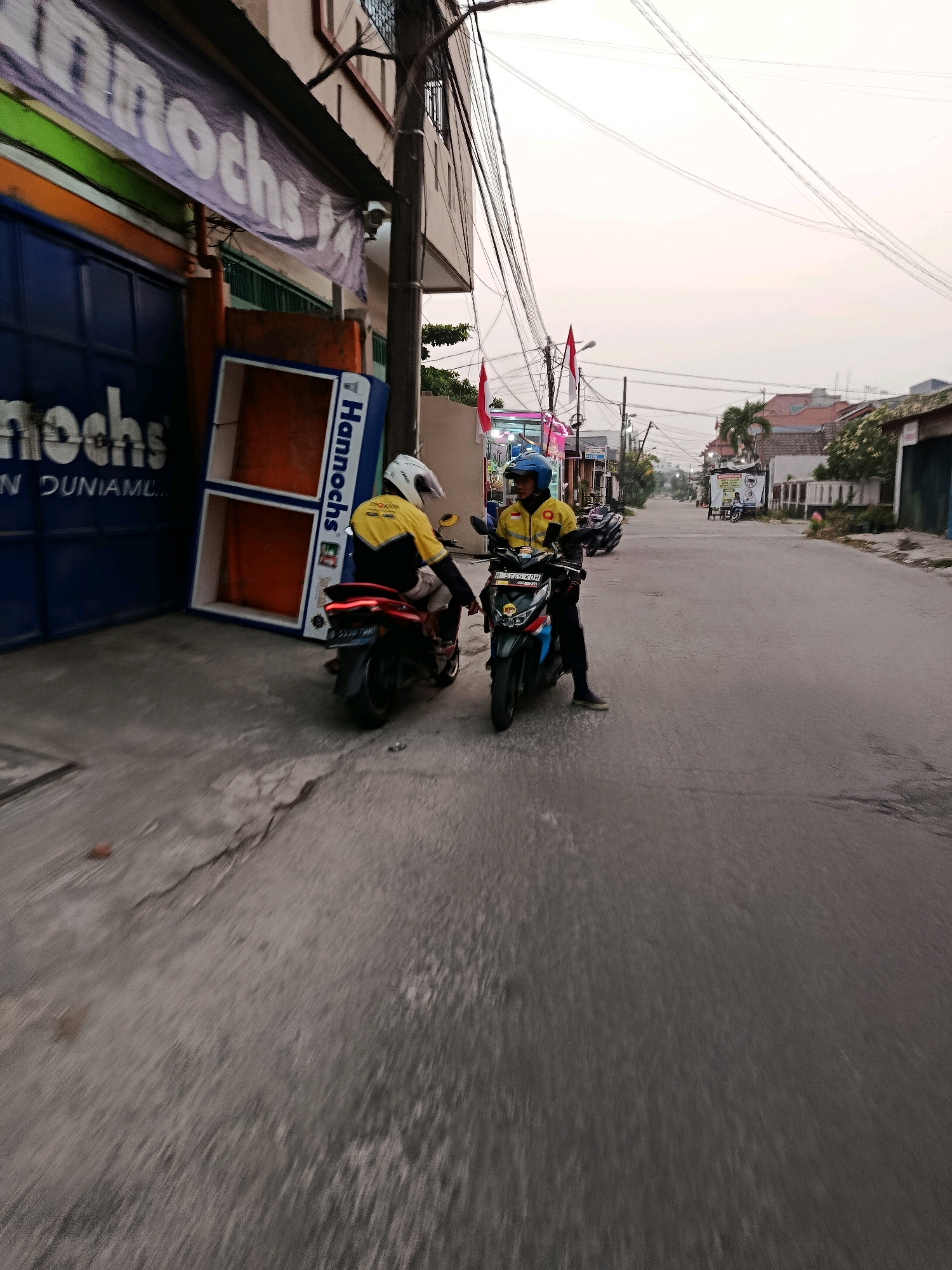 Two individuals wearing yellow jackets and helmets are on motorcycles in front of a building with a blue facade and a purple banner reading "Hannochs" in the city of Bekasi, Indonesia. The building appears to be a commercial establishment. One individual is seated on a red motorcycle, facing away from the camera. The other individual is seated on a dark colored motorcycle, facing the camera. The street is paved and appears to be a residential or commercial area with houses and a few other parked motorcycles visible in the background. The sky is overcast, suggesting it may be early morning or late afternoon. There are electrical wires overhead.