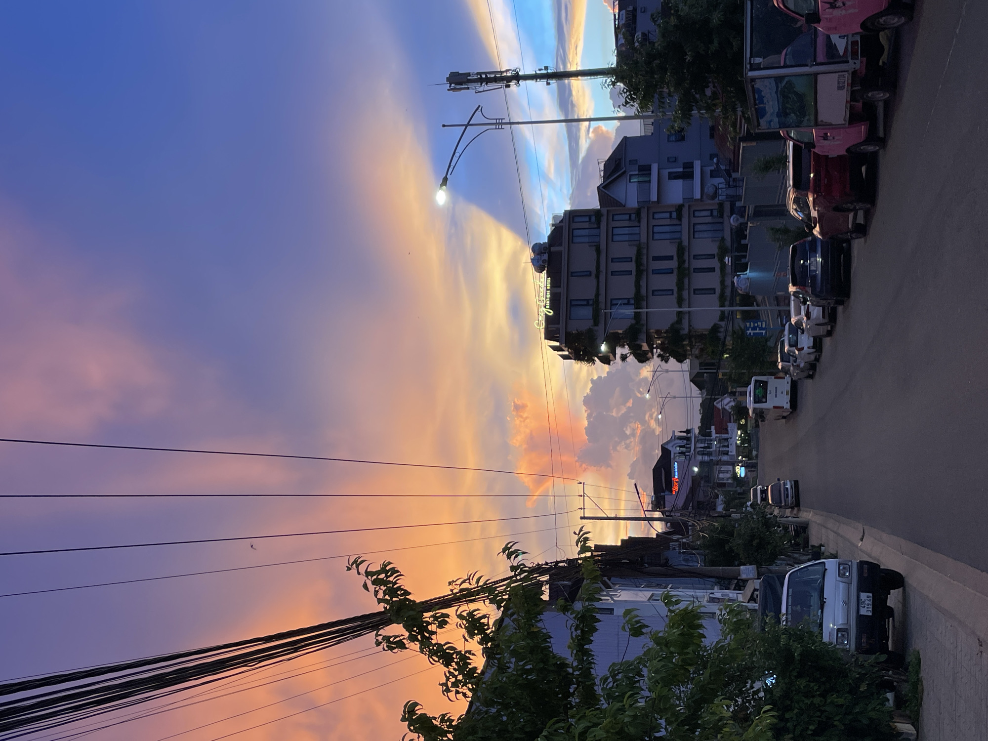 This image depicts a street scene at dusk in Lâm Viên - Đà Lạt, Vietnam. The sky is ablaze with a vibrant sunset, transitioning from deep blue and purple hues at the top to fiery oranges and pinks closer to the horizon. Wispy clouds catch the last rays of sunlight, creating a dramatic and colorful display.
The foreground is dominated by a tangle of power lines, with a street light on one of the poles casting a warm glow. Below, a row of parked vehicles lines the side of a paved road, including several small trucks and cars. The buildings lining the street are a mix of styles, some appearing modern with multiple stories and balconies, while others are more traditional. One building prominently displays a sign that reads "Star Hotel". Green foliage from trees and bushes peeks out from between the buildings and along the sidewalk, adding a touch of nature to the urban setting.
The time of day is clearly dusk, indicated by the sunset colors and the illuminated street light. The overall atmosphere is peaceful and serene, capturing the quiet beauty of a Vietnamese town as day turns into night. There are no people clearly visible in the image.