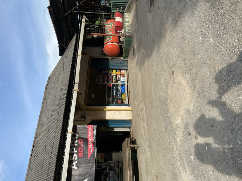 The image shows a storefront in Sukatani, Indonesia (latitude: -6.631, longitude: 107.404) with a sign that reads "ASPRO BOLD Rp 27.000/120 batang". The storefront is made of concrete and has a corrugated metal roof. The storefront has an open doorway and a window displaying various products, including what appears to be bottled beverages and household goods. There is a wooden bench in front of the storefront. A large, red, cylindrical propane tank with a green "UNIPEX" logo is located to the right of the storefront. There are shadows of two people in the foreground. The ground is made of gravel. The sky is blue with some clouds. The environment is a residential or commercial area. There are no people visible in the scene, but the shadows of two individuals are present in the foreground. The scene is set during the daytime, under a clear sky. There are no signs of any activity or interaction occurring at the storefront.