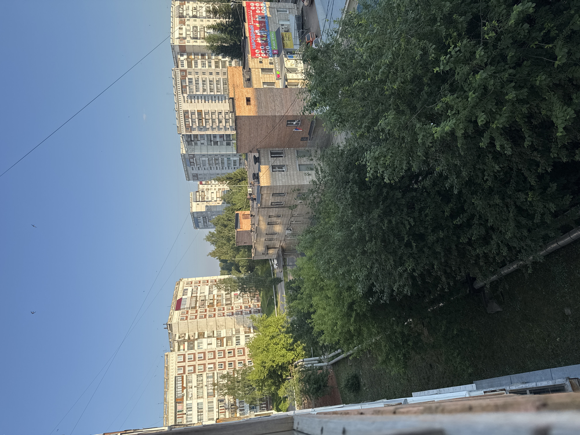 The image captures an aerial view of a residential area, likely in Svetly, Russia, characterized by apartment buildings and lush greenery. In the foreground, the edge of a building with visible pipes suggests an urban environment. Dominating the midground are several multi-story apartment buildings, some with light-colored facades and numerous windows, others appearing older and slightly weathered. A smaller brick building with a prominent red roof element stands out. Across the street, another large apartment complex is visible in the background. The scene is bathed in the warm light of what appears to be late afternoon or early morning. The sky is clear and blue, with a few faint bird silhouettes visible. Trees and dense foliage are interspersed between the buildings and cover the ground, creating a sense of nature within the urban landscape. A white car is parked on the road in the midground. A banner displaying text and graphics is affixed to the side of the brick building.