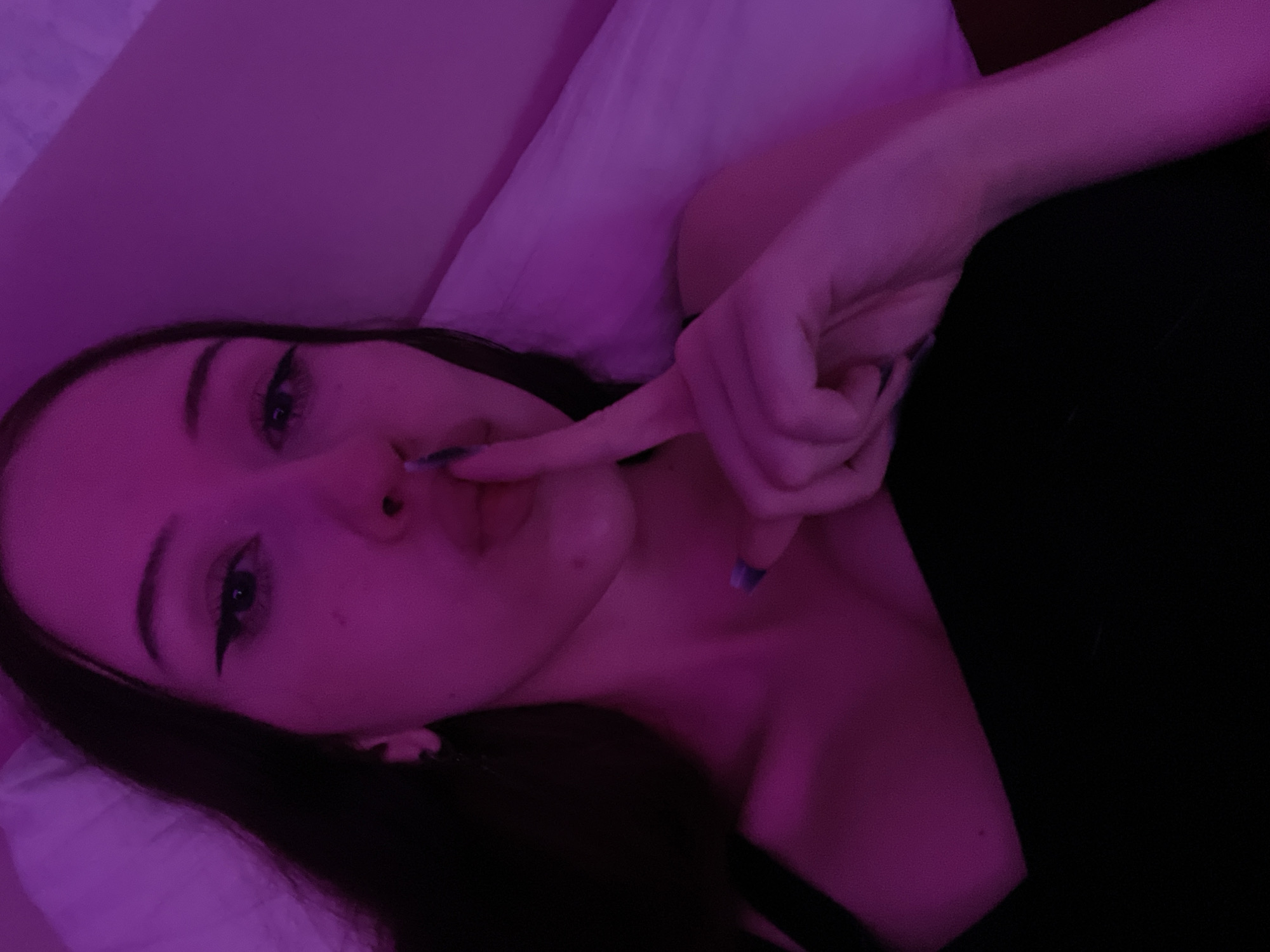 The image is a close-up, low-angle selfie of a young woman in bed, lit by purple ambient light. She is lying on her side, with her head resting on a pillow. Her dark hair is visible, falling around her face and shoulders. She is wearing a black top. Her right hand is raised to her lips, with her index finger pressed against them, in a "shush" gesture. She has eyeliner and her fingernails appear to be painted blue. The background consists of a white pillow and a dark surface, possibly a bedspread or blanket. The overall mood is intimate and secretive. There is no information in the image that suggests a location in Votkinsk, Russia.