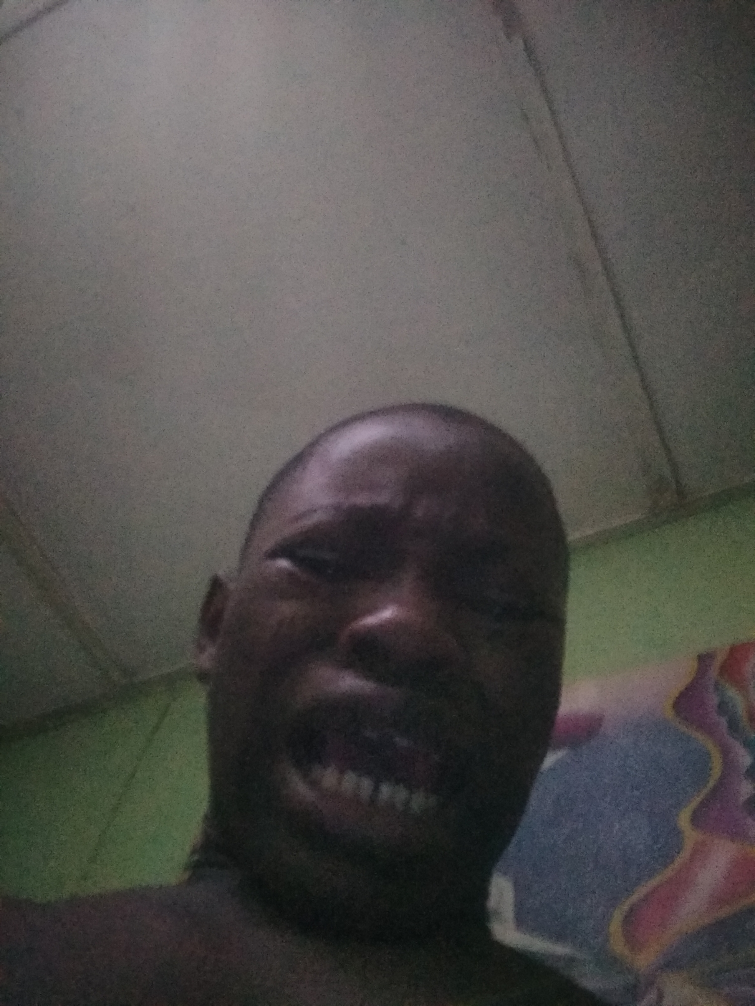 This is a close-up, low-angle selfie of a dark-skinned individual, likely an adult male, taken indoors in a dimly lit environment. The person's face occupies the majority of the frame, captured from below, looking up towards the camera. He appears to have a shaved head or very short hair. His expression is intense, with eyes partially closed or squinting, and his mouth open in a grimace or exaggerated grin, revealing his teeth.
Above the person's head, a light grey ceiling with faint linear indentations is visible. To the left and partially behind his head, there is a wall painted in a muted green color. On the right side of the image, behind the person's shoulder, a section of colorful, patterned fabric, possibly a pillow or bedding, can be seen, featuring shapes in blue, pink, yellow, and white.
The image quality is grainy, suggesting it was taken in low light. There is no visible text. The overall scene depicts a casual, personal moment. The location context provided is Shangisha, Nigeria.