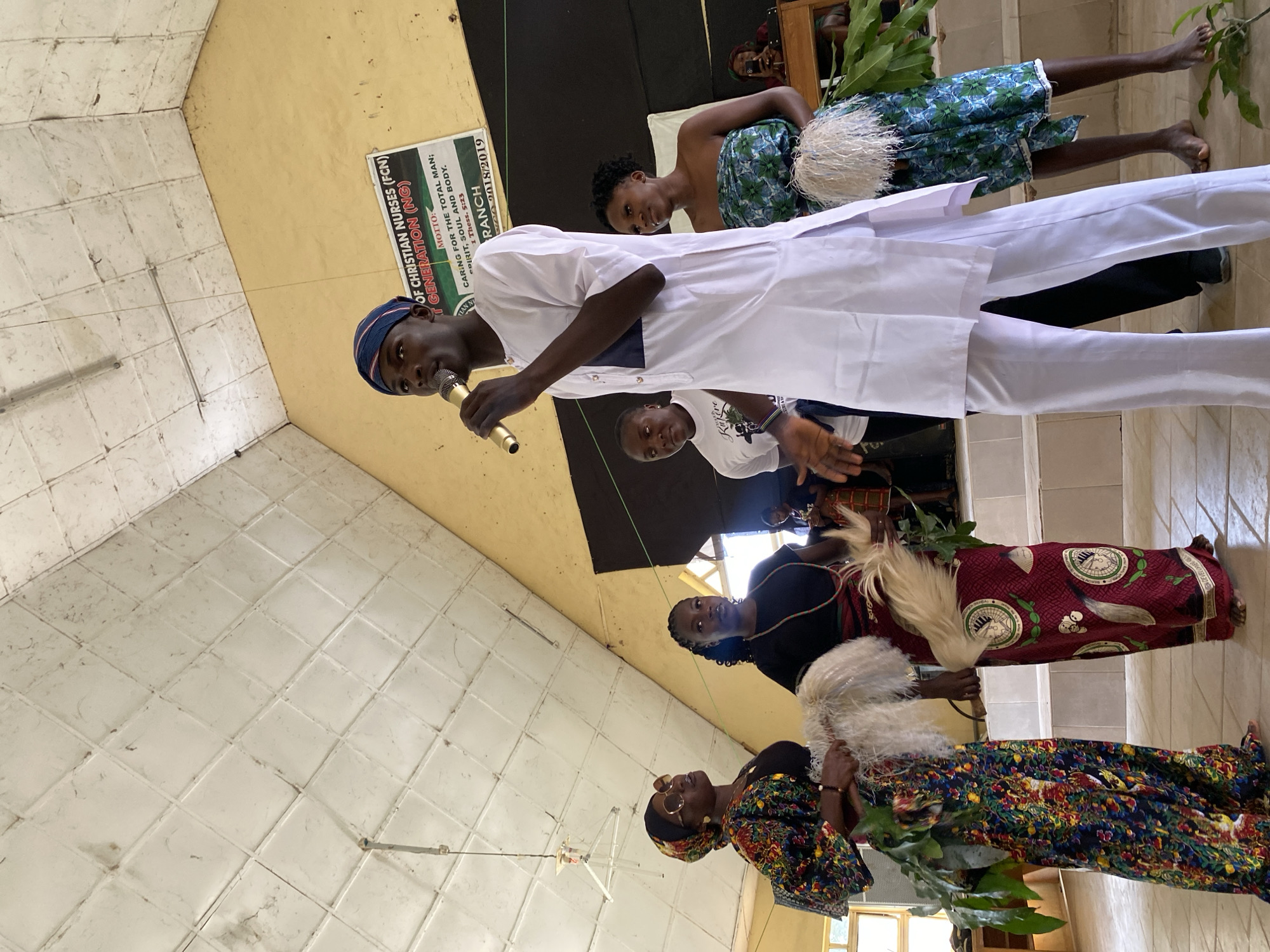 This image captures a gathering, likely a church event, in Dutsen Kato, Nigeria. Several people are present, including a young man speaking into a microphone, dressed in a white traditional outfit and a blue headwrap. He appears to be the focus of attention. Behind him, several women are also visible, some wearing colorful traditional attire. One woman in the background wears a blue and green patterned fabric, while another is dressed in a dark top and a vibrant, patterned skirt. The setting appears to be indoors, with white tiled walls and a yellow ceiling. A banner in the background reads, "OF CHRISTIAN NURSES (FCN) T GENERATION (NG)" with a motto and scriptural reference. The overall atmosphere seems formal yet celebratory, with the presence of traditional garments and what looks like a ceremonial element with white feathery objects held by some of the women. The lighting suggests it could be daytime, with natural light possibly coming from an unseen window. The image is oriented vertically.
