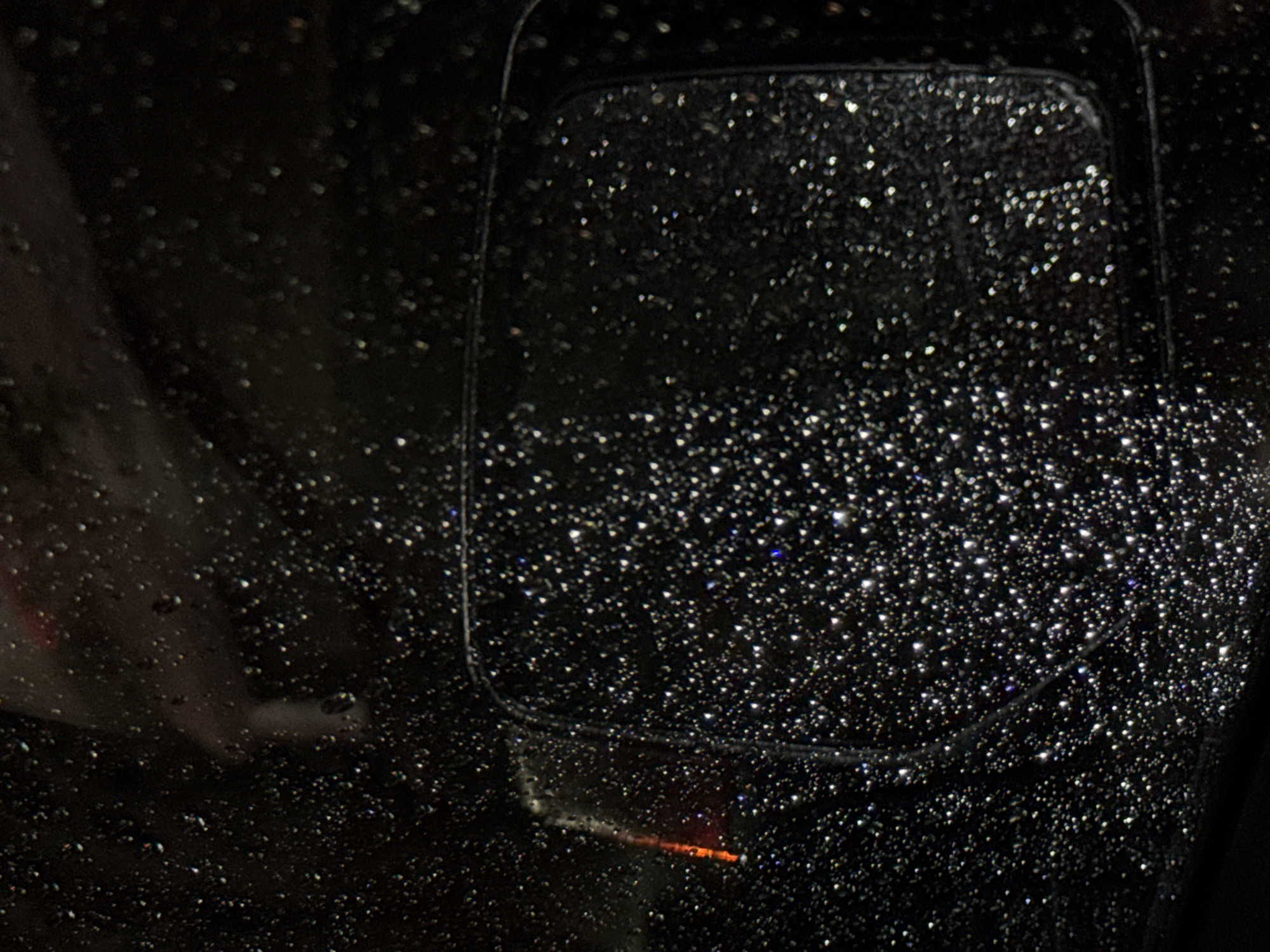 A close-up photograph captures a black vehicle's side mirror heavily covered in water droplets. The mirror itself is a dark, rectangular shape with rounded corners. The water droplets are reflecting light, creating a sparkling effect across the surface of the mirror. The image is taken at night. It's unclear if the location is Mexico City.