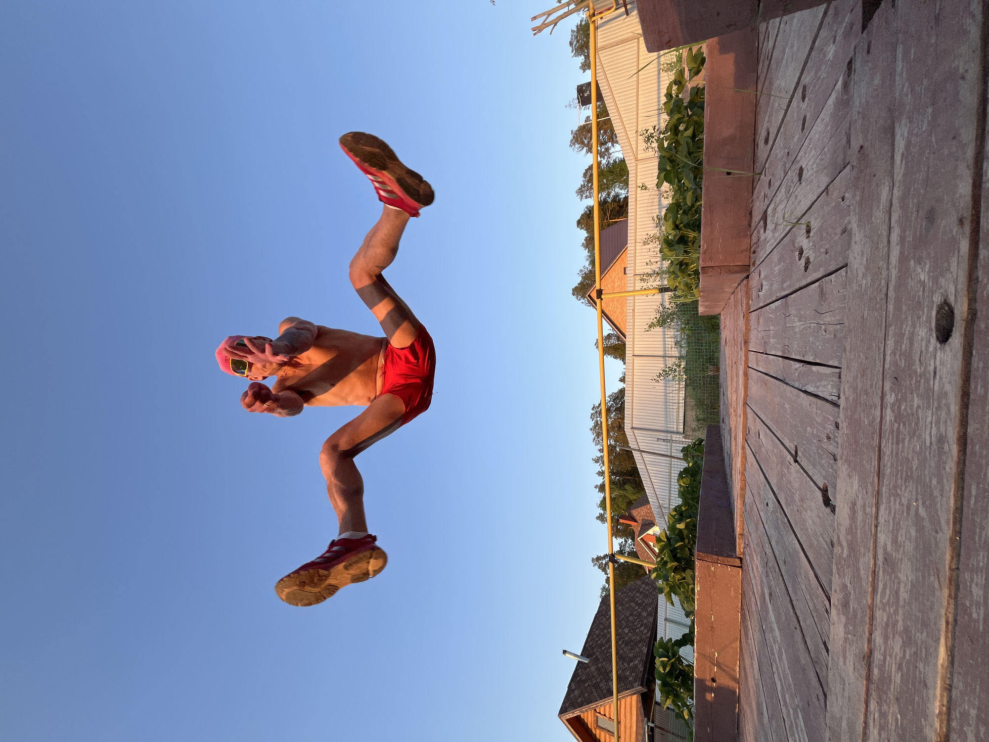 A man in red shorts and red sneakers is in mid-air against a clear blue sky. He's wearing a pink hat and sunglasses, with his arms raised and hands making a gesture. To the right of him is a wooden deck, and beyond that are houses and greenery. The image appears to be taken in a residential area. The location is Chaikovsky, Russia.