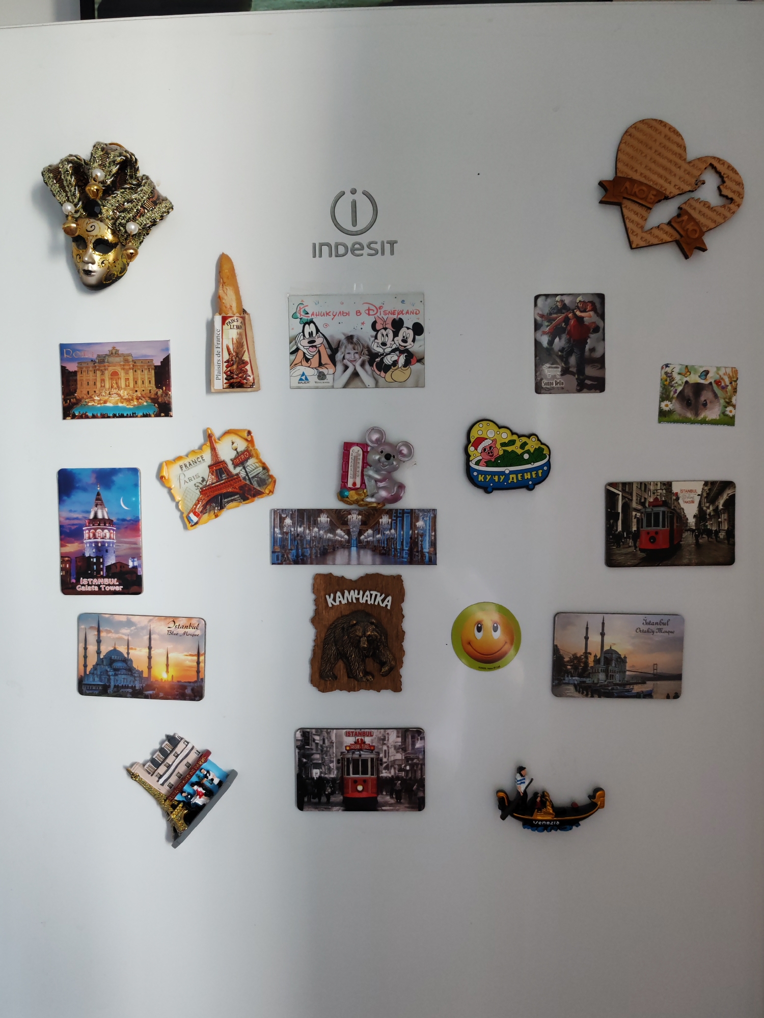A white refrigerator door is covered with a collection of diverse magnets, predominantly souvenirs from various international travel destinations. The scene depicts a personal display of memories and interests, suggesting extensive travel. **Subjects and Objects:** The primary subjects are numerous refrigerator magnets of different shapes, sizes, and materials (wooden, plastic, ceramic, photographic). * **Travel-themed magnets:** These include iconic landmarks such as the Trevi Fountain in Rome ("Rome"), the Eiffel Tower and Arc de Triomphe in Paris ("FRANCE," "ARIS," "METRO," "RESTAURANT DE PARIS"), the Galata Tower and Blue Mosque in Istanbul ("İSTANBUL Galata Tower," "Istanbul Blue Mosque"), an Istanbul tram ("ISTANBUL TAKSİM"), the Ortaköy Mosque in Istanbul ("İstanbul Ortaköy Mosque"), and a gondola from Venice ("Venezia"). There are also magnets dedicated to Disneyland ("КАНИКУЛЫ В DISNEYLAND" - Holidays in Disneyland) featuring Mickey and Minnie Mouse, and several related to Kamchatka, Russia, including a wooden heart-shaped magnet with "КАМЧАТКА" (Kamchatka) and "ЛЮБ" (Lyub), and a wooden magnet with a bear and "КАМЧАТКА". * **Decorative/Miscellaneous magnets:** These include an ornate golden and black Venetian mask, a magnet resembling a baguette ("Plaisirs de France," "PAINS DU LEVAIN"), a grey mouse holding a thermometer, a green smiling face, a pig in a bathtub with "КУЧУ ДЕНЕГ" (A lot of money), a small magnet featuring a hamster among flowers, and a magnet depicting two men in a dramatic scene with "Sonno Bello" text. * **Branding:** The refrigerator itself is an "INDESIT" appliance. **Activity or Event:** There is no direct activity or event depicted other than the static display of magnets. The collection implies the activity of travel and souvenir collecting. **Notable Details:** The diverse origins of the magnets suggest the owner has traveled extensively, with a particular interest in France, Italy, Turkey, and Kamchatka in Russia. The presence of text in English, French, and Russian further supports this. The items are personal mementos. **Visible Text:** * "INDESIT" (refrigerator brand) * "Rome" * "Plaisirs de France" * "PAINS DU LEVAIN" * "КАНИКУЛЫ В DISNEYLAND" (Holidays in Disneyland) * "Sonno Bello" * "İSTANBUL Galata Tower" * "FRANCE" * "ARIS" * "METRO" * "RESTAURACE DE PARIS" (appears on two separate Paris magnets) * "КУЧУ ДЕНЕГ" (A lot of money) * "ISTANBUL TAKSİM" * "Istanbul Blue Mosque" * "КАМЧАТКА" (repeated on wooden heart magnet and a bear magnet) * "ЛЮБ" (on wooden heart magnet) * "Venezia" * "İstanbul Ortaköy Mosque" The variety of magnets indicates a resident of Saint Petersburg,