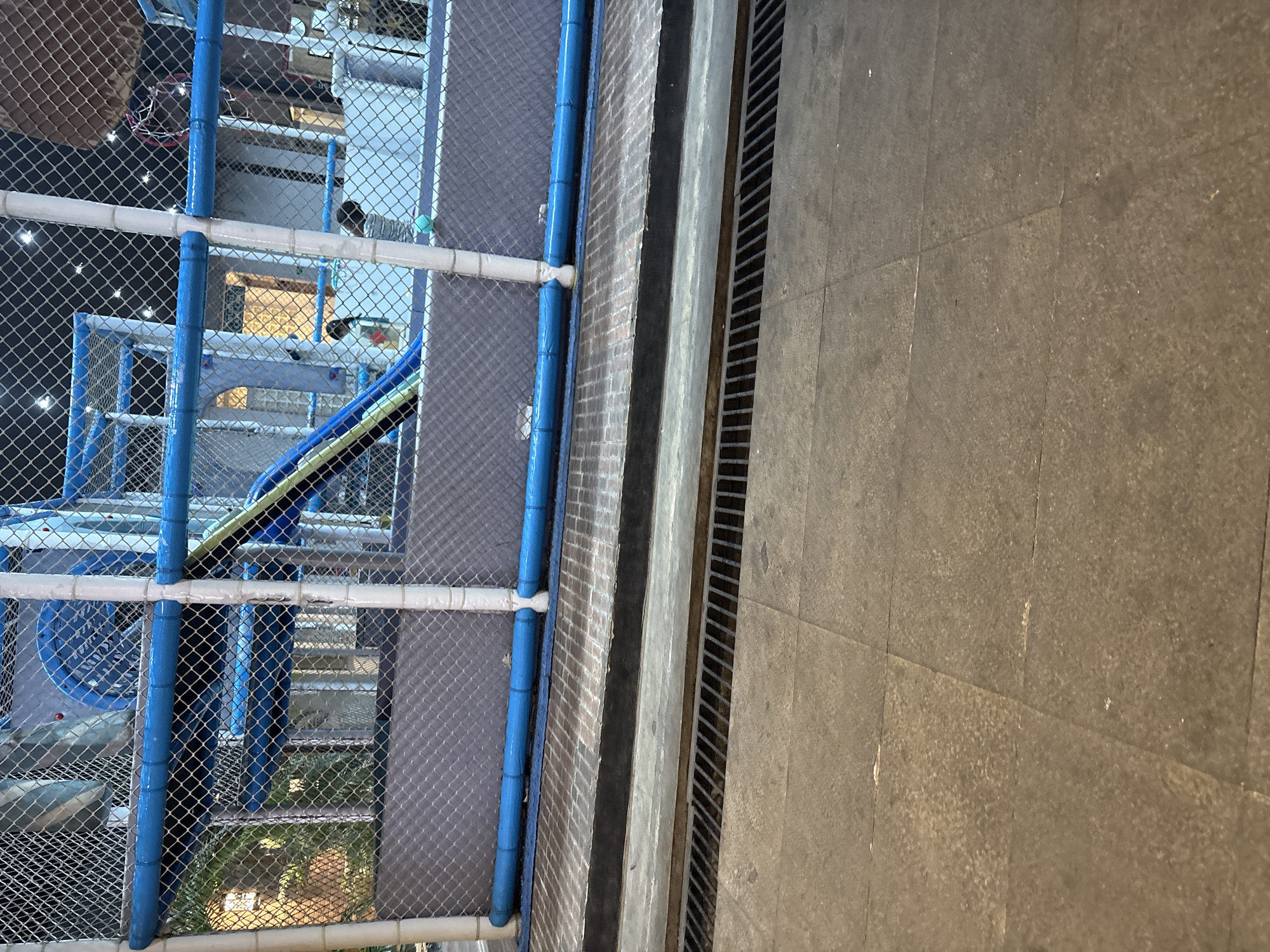 The image depicts an indoor play area, possibly a children's entertainment center, located in Cirebon, Indonesia. In the foreground, a metal grate runs horizontally across the scene, suggesting a drainage system. The grate is positioned in front of a raised platform covered with dark gray padding. This platform is bordered by a bright blue pipe that runs along its length, supported by white cylindrical posts. Behind this, a mesh fence encloses the play structure, which features blue slides, netted tunnels, and white support poles. Several individuals, including children, are visible within the play area, engaged in various activities, though their actions are partially obscured. The lighting suggests the photo was taken indoors, likely during evening hours, with artificial lights casting a warm glow on the structures.