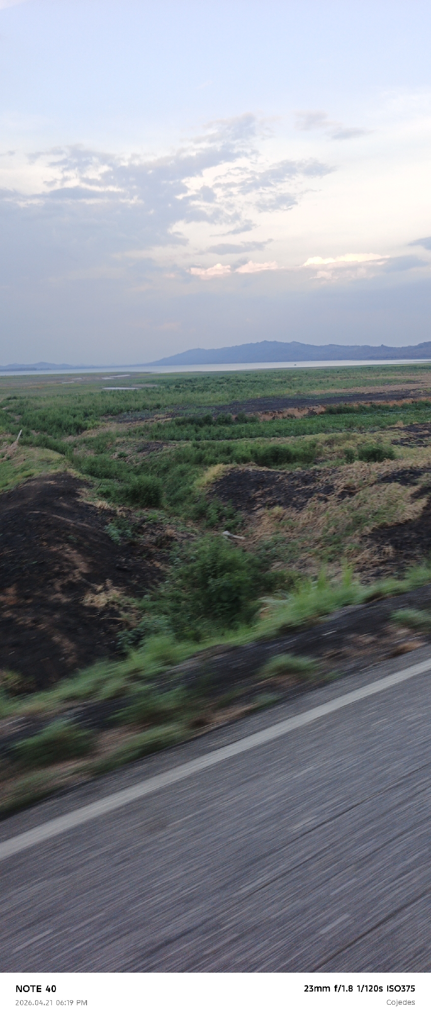 The image shows a view from a moving vehicle, likely on a road near a body of water. In the background, there's a lake or reservoir with land extending into the water, possibly a peninsula or low-lying area. Beyond the water, a mountain range is visible. The sky is overcast with clouds. The middle ground features a landscape with varying textures, including patches of green vegetation interspersed with areas of dark, possibly burnt ground. In the foreground, the road surface is visible, blurred due to motion. The image includes camera settings and location information at the bottom. The location indicated is Cojedes, which is a state in Venezuela. The city listed is La Galera in the Cojedes.
