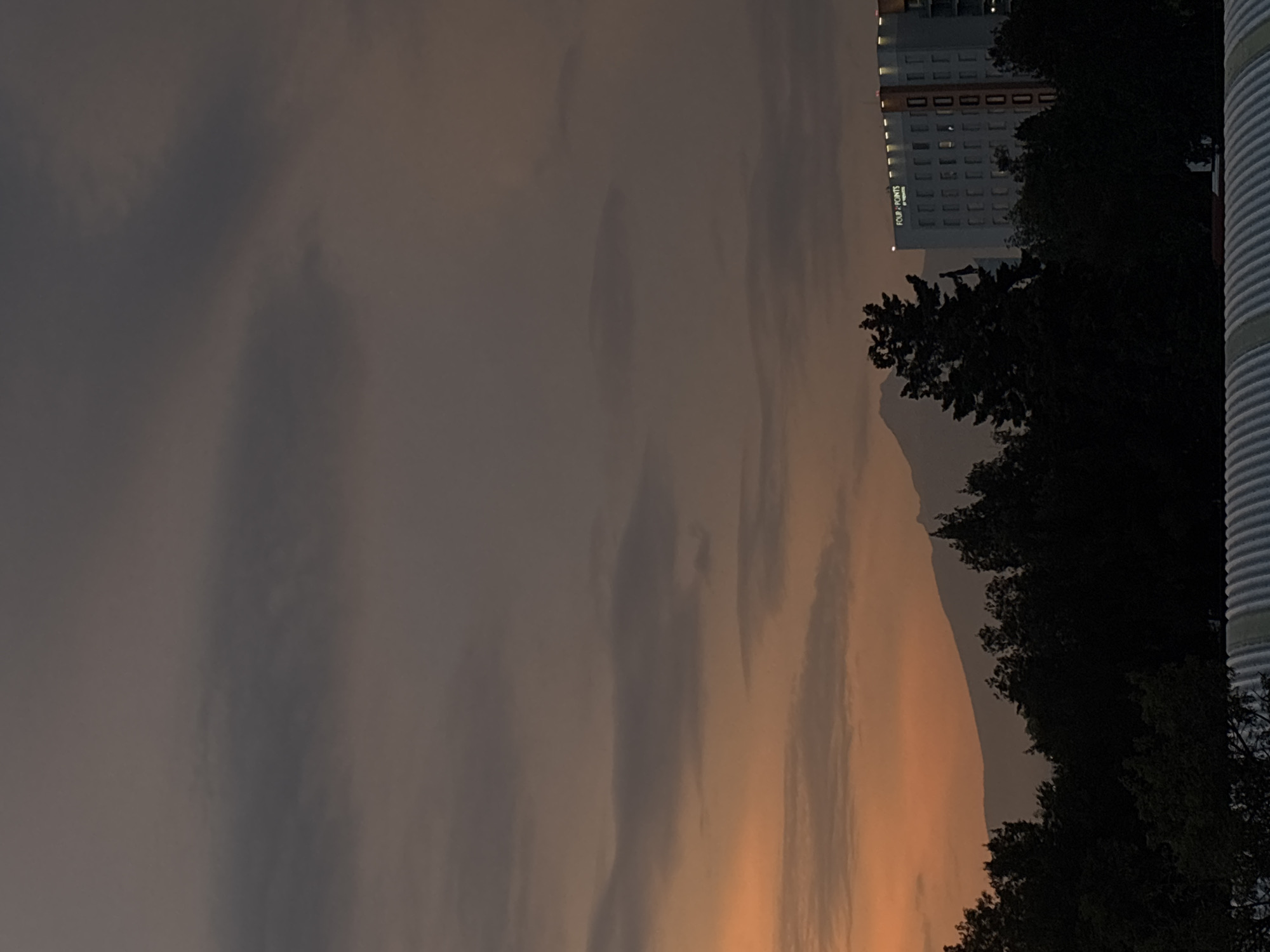 The image depicts an evening scene, possibly in Puebla, Mexico, suggested by the presence of a building with "FOUR POINTS BY SHERATON" signage, which is a hotel located in Puebla. The sky transitions from a warm, orange hue near the horizon to a greyish tone higher up, indicative of sunset or twilight. A mountain silhouette is visible in the middle ground, partially obscured by trees in the foreground. A tall building is visible to the upper right of the mountain, and a corrugated metal structure is to the right side. The general impression is of a cityscape viewed during a colorful sunset. There are no people visible.