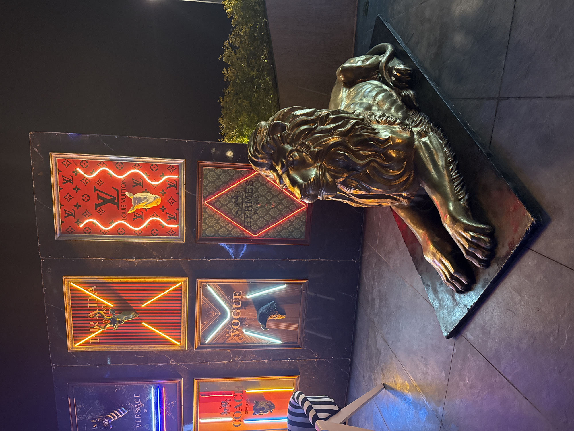 The image shows an outdoor display in Kemer, Turkey. A large, bronze-colored lion statue rests on a dark stone base. Behind it is a panel with framed brand logos including Louis Vuitton, Hermes, Prada, Vogue, Versace, and Coach, all illuminated with neon lights. The background includes a dark sky and some foliage. The scene is captured at night.