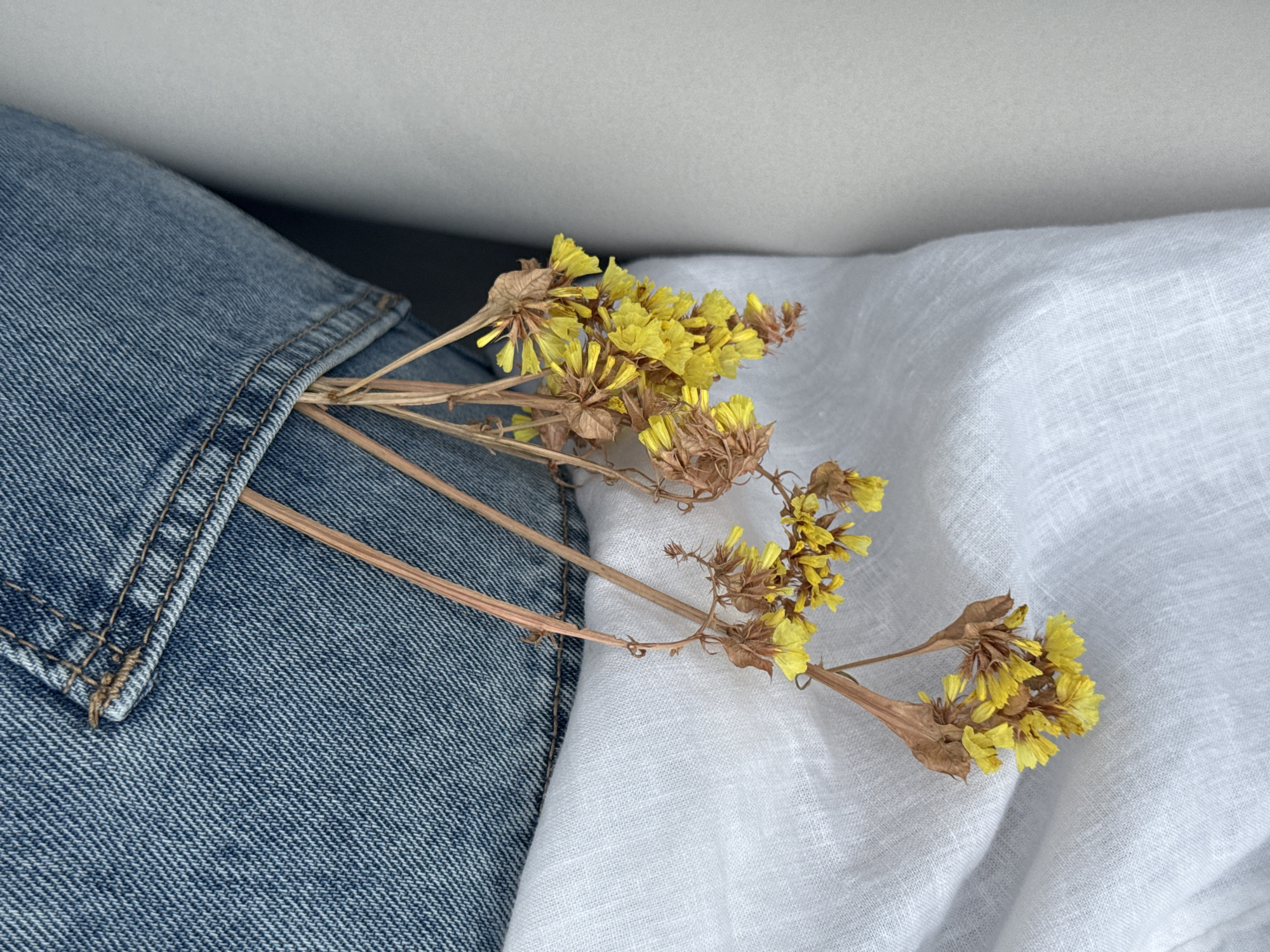A close-up shot shows a bouquet of dried yellow flowers placed across a light blue denim jacket and a white linen fabric. The flowers are positioned with their stems tucked into the jacket's pocket. The background is a solid gray, suggesting an indoor setting. No people are visible in the frame. There is no evidence to suggest the location is Chaikovsky, Russia.