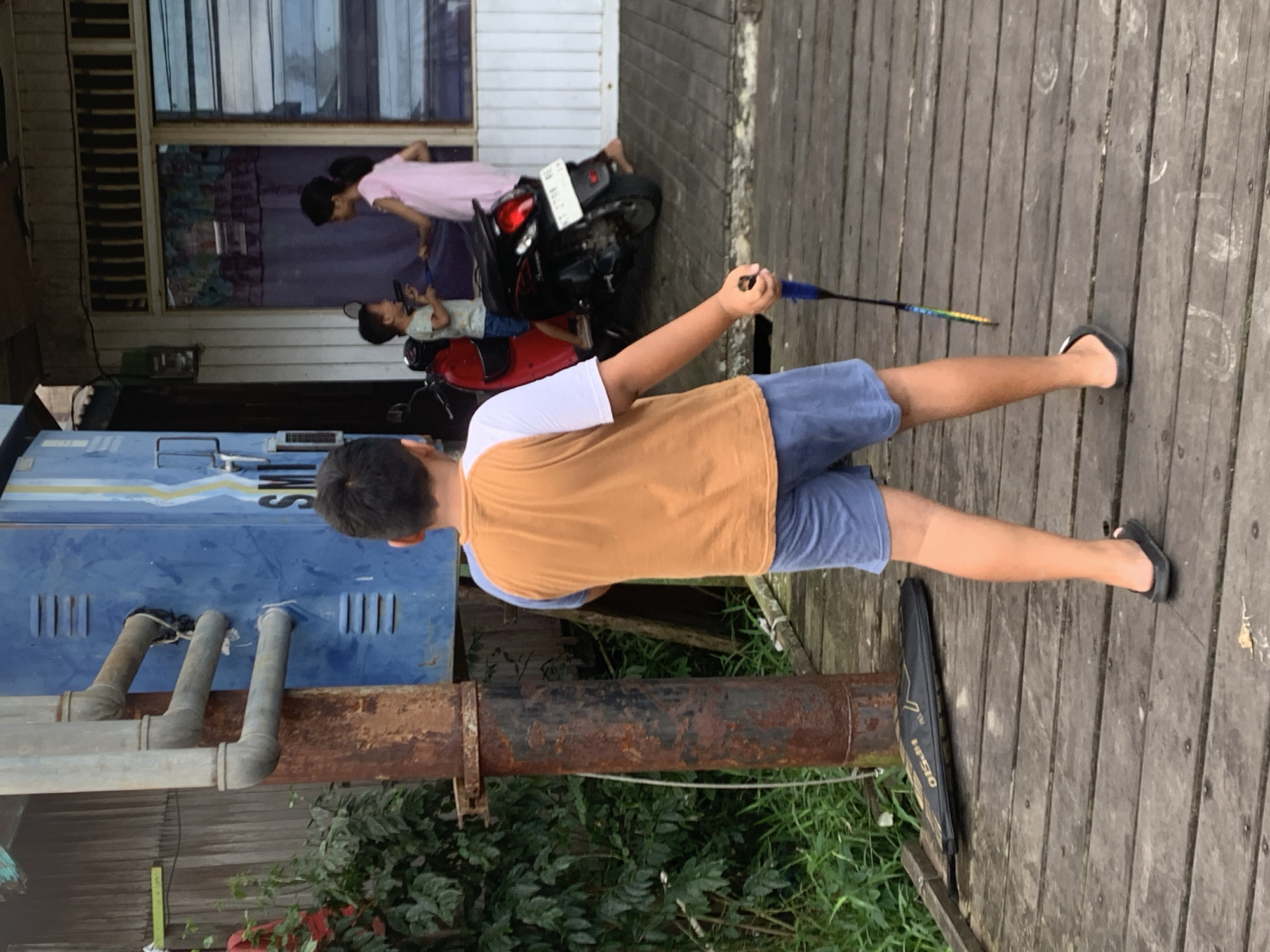 This media file presents an outdoor daytime scene set on a wooden deck in Sungai Meriam, Indonesia. In the foreground, a young person, viewed from behind, wears a white t-shirt with an orange-brown layer, blue shorts, and black flip-flops. They are holding a colorful badminton racket in their right hand, suggesting activity or preparation for play. A black carrying case, possibly for sports equipment, with "HPS10" visible on its side, rests on the wooden planks of the deck beside them.
Further into the midground, a young girl in a pink top is seen interacting with a smaller child who is seated on a red scooter. Behind them, a black motorcycle is parked. These figures are positioned in front of a building featuring windows and horizontal wooden slats in its structure.
To the left of the foreground subject, a blue utility box or container is visible, from which three gray pipes extend downwards. Partial text "SMT" is discernible on this blue structure. A large, rusted metal pipe runs vertically below, while lush green foliage and plants grow at the ground level beneath the elevated wooden platform. The natural lighting indicates it is daytime, and the overall atmosphere is casual.