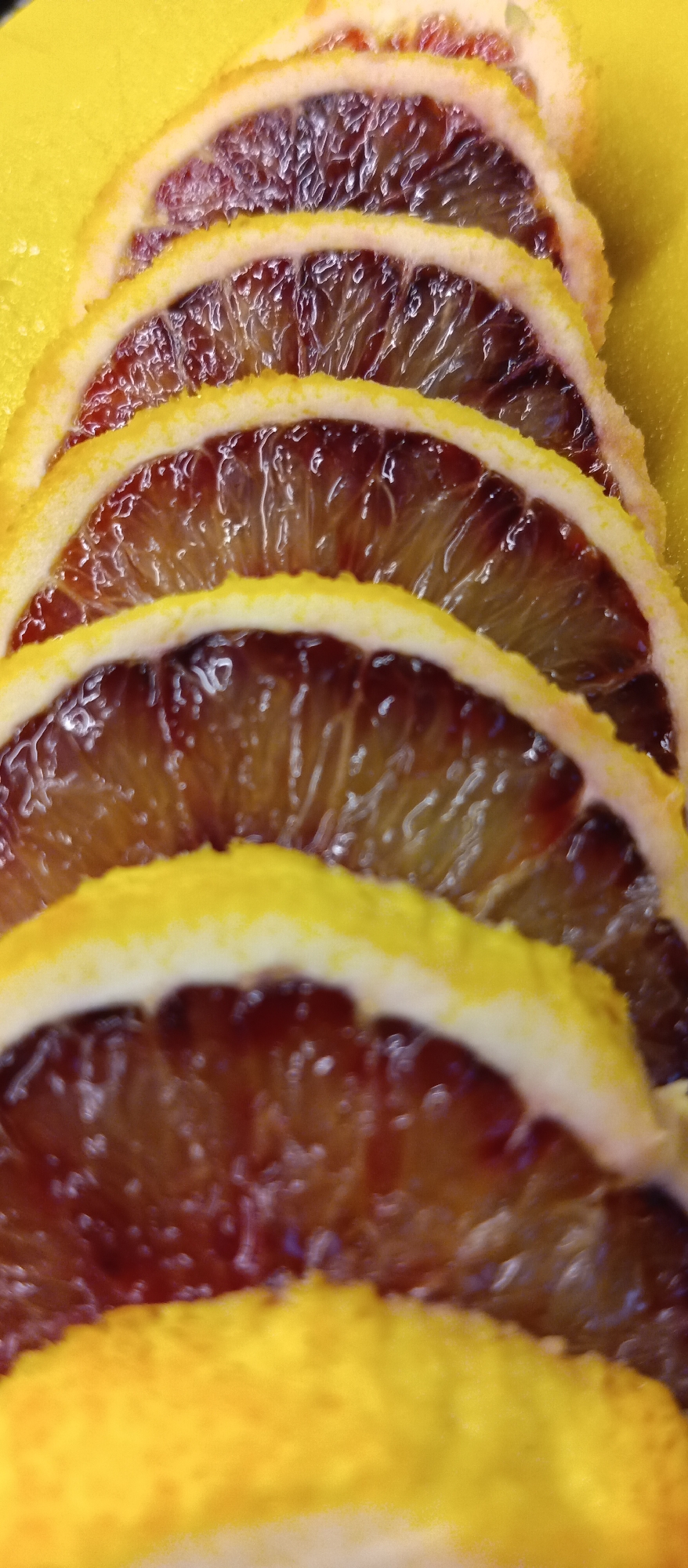 The image shows a close-up of several slices of a blood orange arranged in a row. The slices are semi-circular, with a thick yellow rind and dark red flesh. The flesh appears juicy and translucent, with visible segments. The background is a solid yellow color. The image does not appear to be located in any specific city.