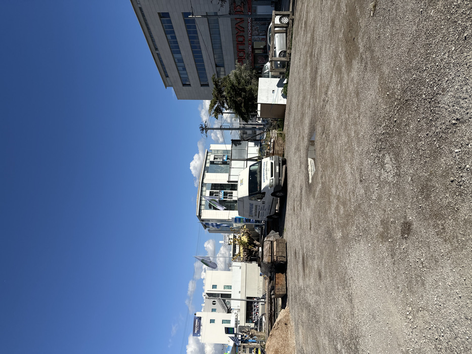 This image, presented on its side (rotated 90 degrees clockwise), depicts a bright, sunny daytime scene in Metamorfosi, Greece. The foreground is dominated by a large, unpaved gravel or dirt lot.
In the mid-ground, a white commercial van is parked at the edge of this lot, facing towards the right. The van has visible Greek text, identifying it as "ΜΟΥΜΟΥΤΣΕΛΗΣ" (Moumoutselis) and indicating services like "ΔΟΜΙΚΑ ΥΛΙΚΑ" (Building Materials) and "ΘΕΡΜΑΝΣΗ-ΨΥΞΗ" (Heating-Cooling), along with contact phone numbers. Adjacent to the van, stacks of materials, possibly building supplies or wooden pallets, are visible.
Beyond the lot and van, a street or road is lined with various buildings. In the upper left background, a multi-story, light-colored building with multiple windows is prominent. Further into the scene, other commercial structures are visible. One building displays a partial sign with Greek text, including "ΛΟΥΛΑ" and "ΕΤΟΝ ? ΤΡΟΦΙΜΩΝ" (Eton ? Foodstuffs). Another building features a blue and white flag or banner, possibly a Greek flag or corporate logo. A sign reading "VICTORIA" is visible on a building further down the street. Power lines and poles are scattered throughout the built environment.
The sky is a clear, vibrant blue with scattered white clouds, indicating sunny weather. The strong sunlight casts distinct shadows, suggesting the time of day is mid-morning to afternoon. No people are visible, and the scene appears quiet and still. The setting is consistent with a commercial or industrial area.