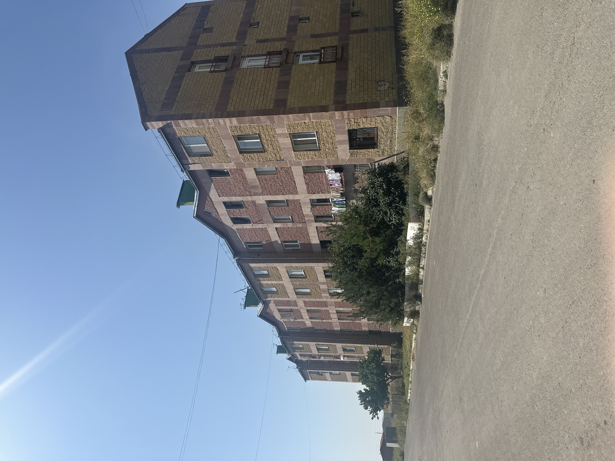 A multi-story apartment building with a facade of yellow and red brick is situated on the left side of a paved road under a clear blue sky. The building has a stepped roofline and features numerous windows and balconies. A line of laundry is hanging from a balcony on the left side of the building. Green trees and bushes are planted in front of the building, and there is dry grass and other vegetation along the roadside. In the distance, there are other structures, including a small building with a red roof. The scene appears to be in a somewhat rural or semi-urban area. This is likely a Soviet-era apartment block in Gyumri, Armenia, a city known for its distinctive architecture and history of seismic activity.
