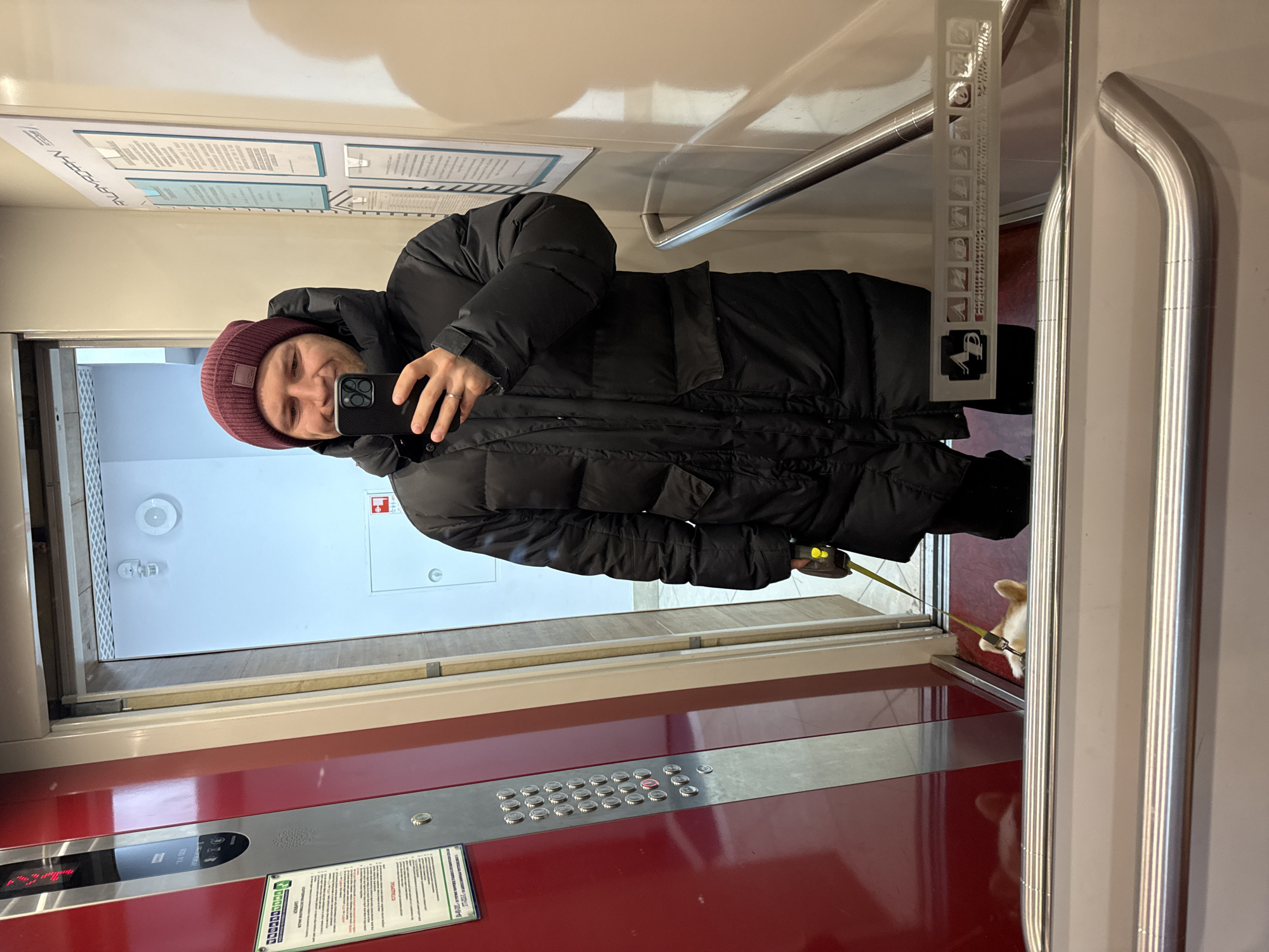 A male individual is captured in a mirror selfie within an elevator in Krasnoyarsk, Russia. He is smiling and holding a smartphone, with which he is taking the picture. He is wearing a dark black puffer coat and a maroon knit beanie, which has a small white logo on it (possibly "SPOON" or similar). A silver ring is visible on his left hand.
The elevator interior features red and silver panels on the walls. A control panel on the red wall displays numerous silver buttons, with one red illuminated button, and a digital display showing the number "2" and an upward arrow, indicating the current floor or destination. Below the control panel, a sign in Russian is visible, titled "ВНИМАНИЕ" (Attention) and "ПРАВИЛА ПОЛЬЗОВАНИЯ ЛИФТОМ" (Rules for using the elevator), along with smaller text and a green and white sticker with a phone number.
In the bottom right of the frame, partially visible behind the elevator door frame, is a small light-colored dog on a yellow leash, suggesting it is accompanying the person. Polished metal handrails run along the elevator walls. Reflected in the mirror, outside the elevator, is a hallway with light-colored walls and several framed documents or posters, some containing text that is difficult to read due to reflection and angle, but one partially shows "A.L.A.V.Z.H.R.A.N" (mirrored). A clear vertical strip on the wall near the handrail shows various pictograms, including a person with luggage, a baby stroller, and a dog, beneath the partially visible Russian text "ЭВАКУАЦИОННАЯ СХЕМА" (Evacuation scheme). The overall scene suggests a casual moment while traveling in an elevator with a pet.