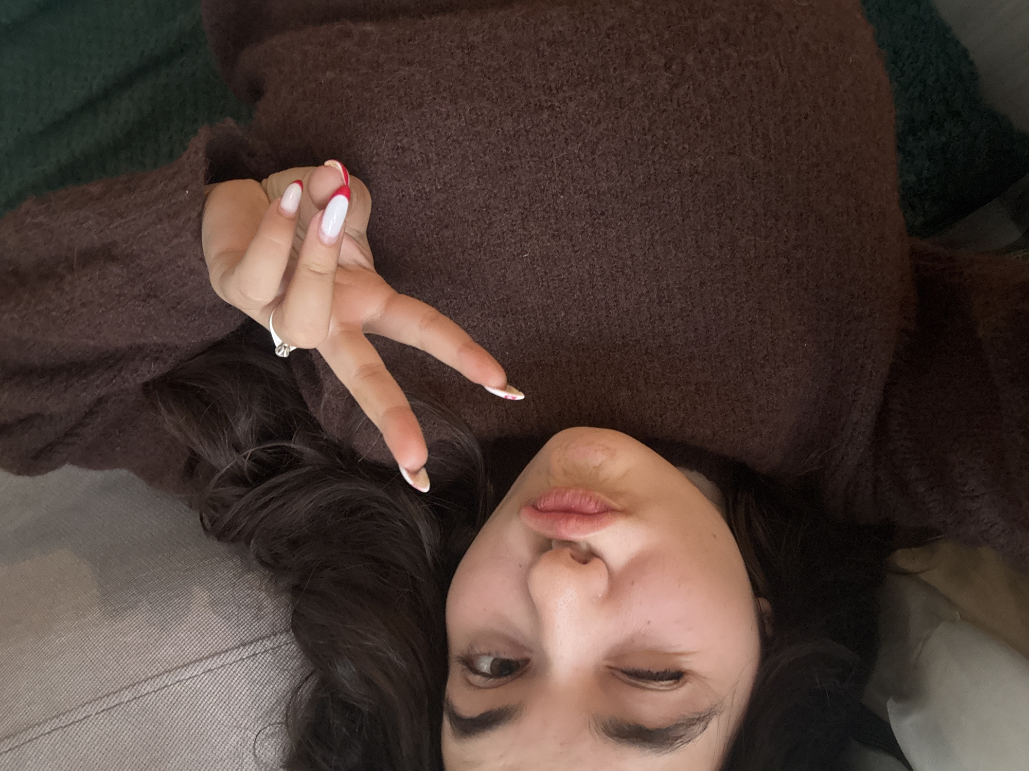 A woman with dark hair and fair skin lies on a gray surface with a brown sweater. She is making a peace sign with her fingers. Her nails are white with a red tip. She is winking with one eye and puckering her lips. The location is not apparent from the image, so I cannot provide an accurate description of the city of Plovdiv, Bulgaria.