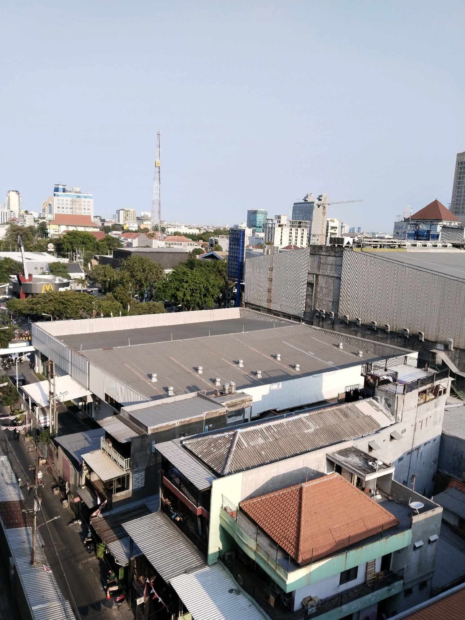 This aerial photograph captures an urban landscape in Surabaya, Indonesia. The scene is dominated by a dense collection of buildings, ranging from multi-story commercial structures to smaller, more densely packed residential or shop-like buildings. In the foreground, large, flat-roofed commercial buildings with metal sheeting are prominent. Moving further into the scene, the architecture becomes more varied, with a prominent red-tiled pyramid-shaped roof on a lighter colored building. Numerous trees are interspersed among the buildings, indicating pockets of green space within the urban sprawl. A tall communication tower is visible in the distance, along with several other high-rise buildings characteristic of a city center. The sky is clear and a pale blue, suggesting fair weather. The streets below are visible in some areas, with some evidence of vehicle and pedestrian activity, though details are indistinct from this vantage point. The overall impression is one of a bustling, built-up urban environment.