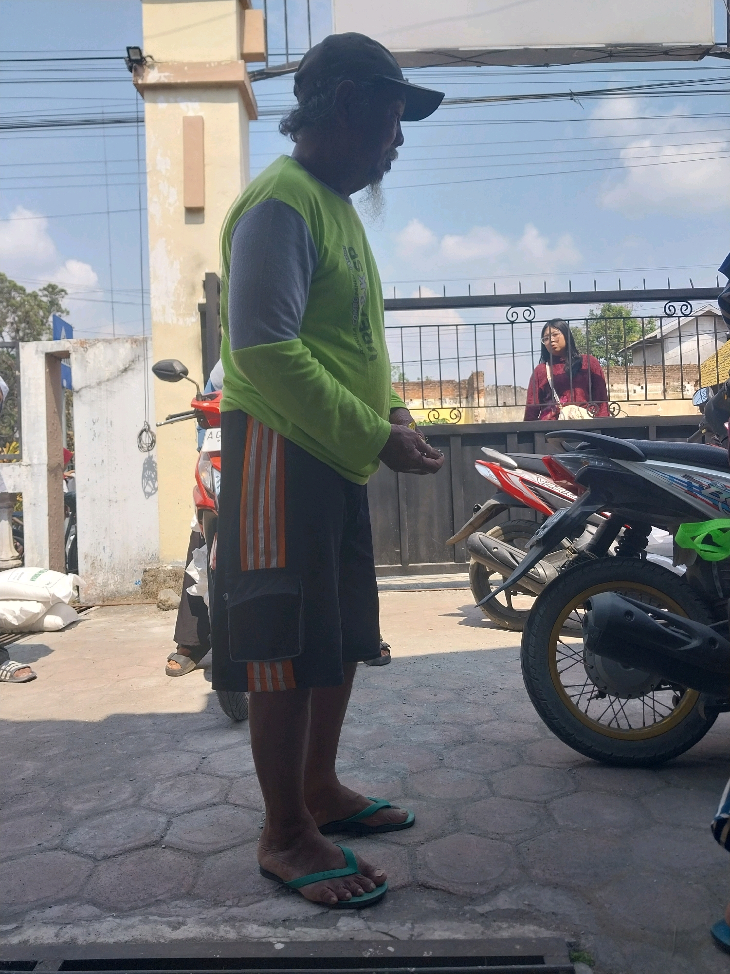 A man wearing a green shirt with gray sleeves, black shorts with orange stripes, and green flip-flops stands on a hexagonal paved walkway in front of a building. He has a beard and wears a dark baseball cap. To his left are sacks of white material and parts of motorcycles. Behind him is a black metal fence, and beyond that, a young woman wearing a maroon sweater and glasses stands looking towards the viewer. The background shows a bright sky with clouds, trees, and buildings. The location is identified as Nglegok, Indonesia.