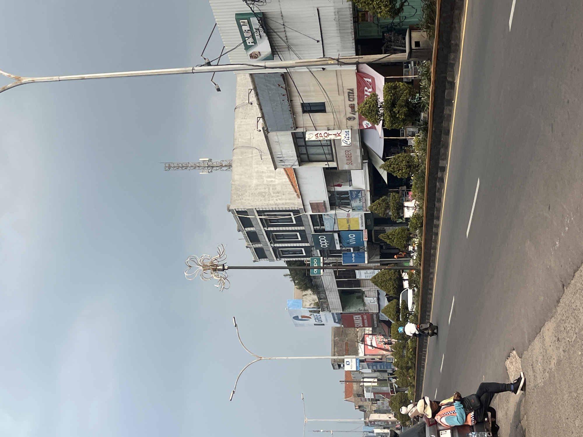 The image shows a street scene in Cilegon, Indonesia, with a focus on a row of buildings lining the right side of a road. The sky is overcast. A person is sitting on the back of a parked vehicle on the left side of the road, wearing a hat and a brightly colored vest over a dark shirt and pants. The person is looking at a phone. A motorcycle with a rider wearing a blue helmet is driving on the road. The buildings on the right are a mix of styles and heights, with several storefronts visible. Signs for various businesses, including "OPPO," "VIVO," "GREE," and "FUJIFILM," are displayed on the buildings. Some of the storefronts have awnings. Trees and shrubbery are planted along the median and sidewalk. A tall street lamp with a decorative top is visible in the foreground, as well as a telecommunications tower in the background. The road is paved with asphalt and marked with white lines.