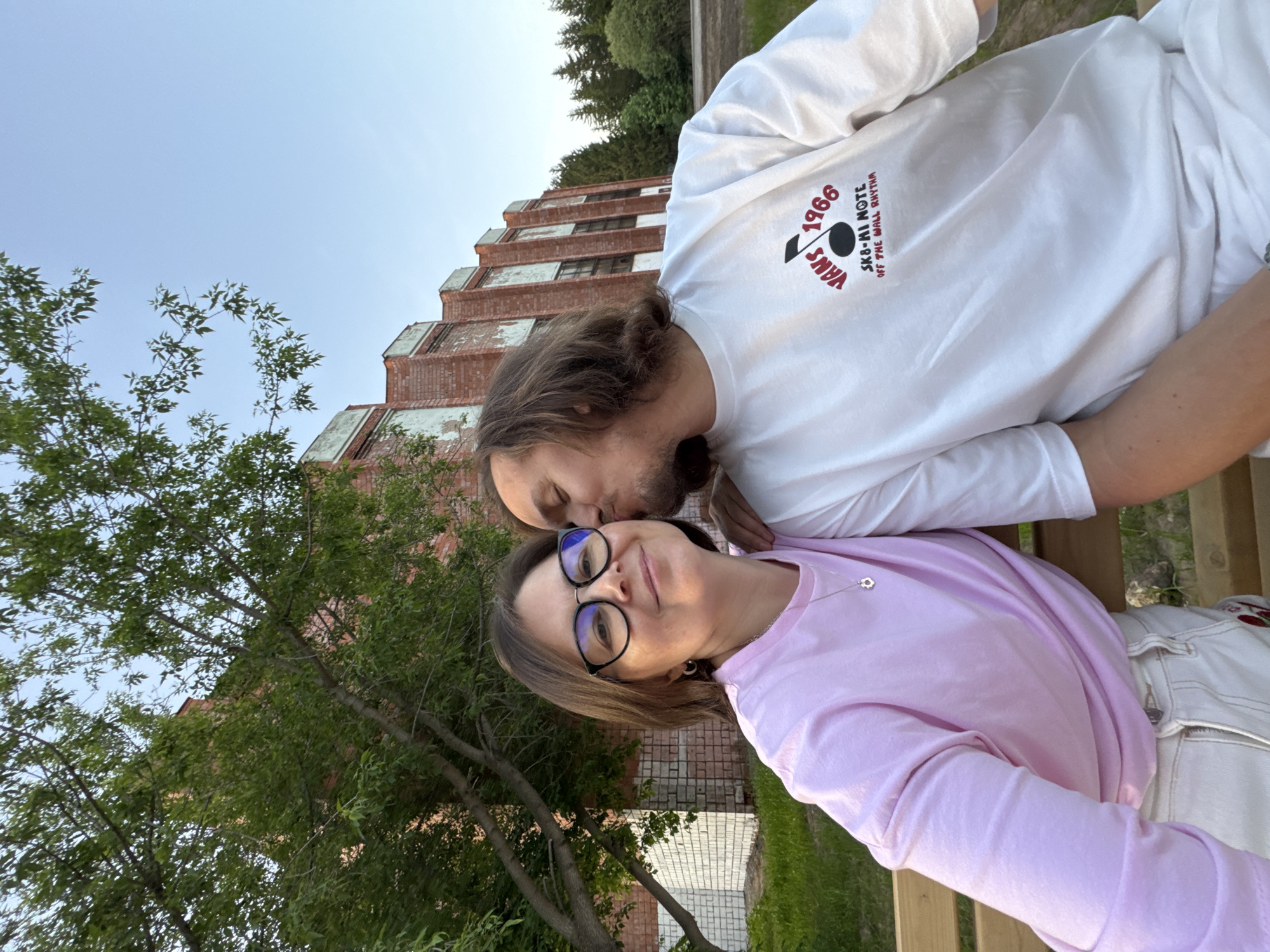 A man and woman are posing for a photograph outdoors, with an urban setting visible in the background. The man, with long brown hair, is wearing a white long-sleeved shirt with Vans branding. He is kissing the woman on the cheek. The woman has brown hair and is wearing glasses and a pink long-sleeved shirt.
Behind them is a brick building, and trees are visible in the background. The location is in Izhevsk, Russia.