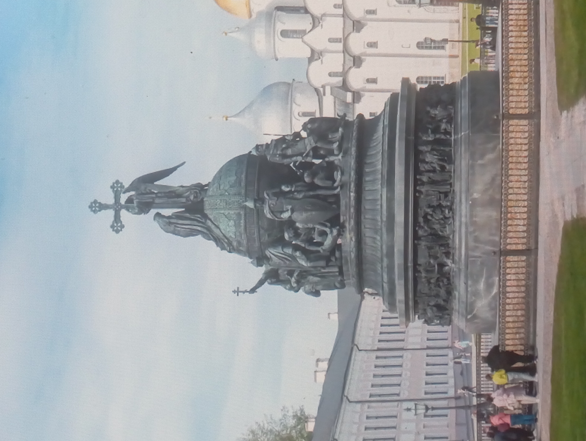 The image depicts a large monument, likely bronze, featuring numerous figures and a cross with an eagle on top. The monument is located in front of several white buildings with domes, possibly churches or cathedrals. A fence borders the monument, and people are standing nearby on a paved area with a grassy patch. The sky is light blue. The image does not contain the city of Karaganda, Kazakhstan. It is likely located in Russia.