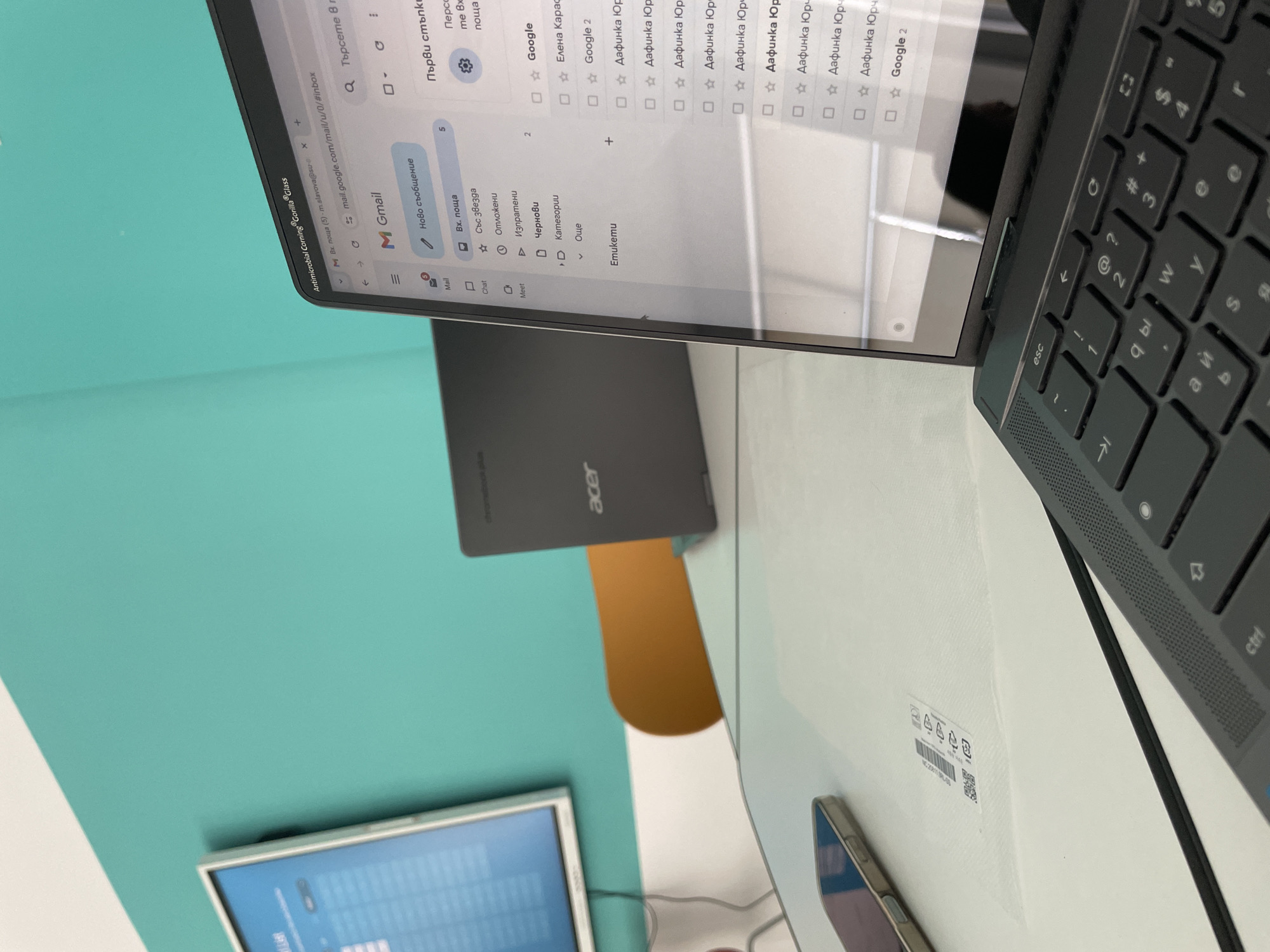 The image shows a laptop displaying a Gmail inbox with a few emails from "Дафинка Юр." A smartphone is positioned near the laptop on a white surface that is likely a desk or table. An Acer Chromebook is nearby, and a monitor is visible in the lower-left. The keyboard is black and features Cyrillic letters. The surrounding environment is a teal-colored wall. It is not possible to confirm that this image was taken in Zlatograd, Bulgaria.